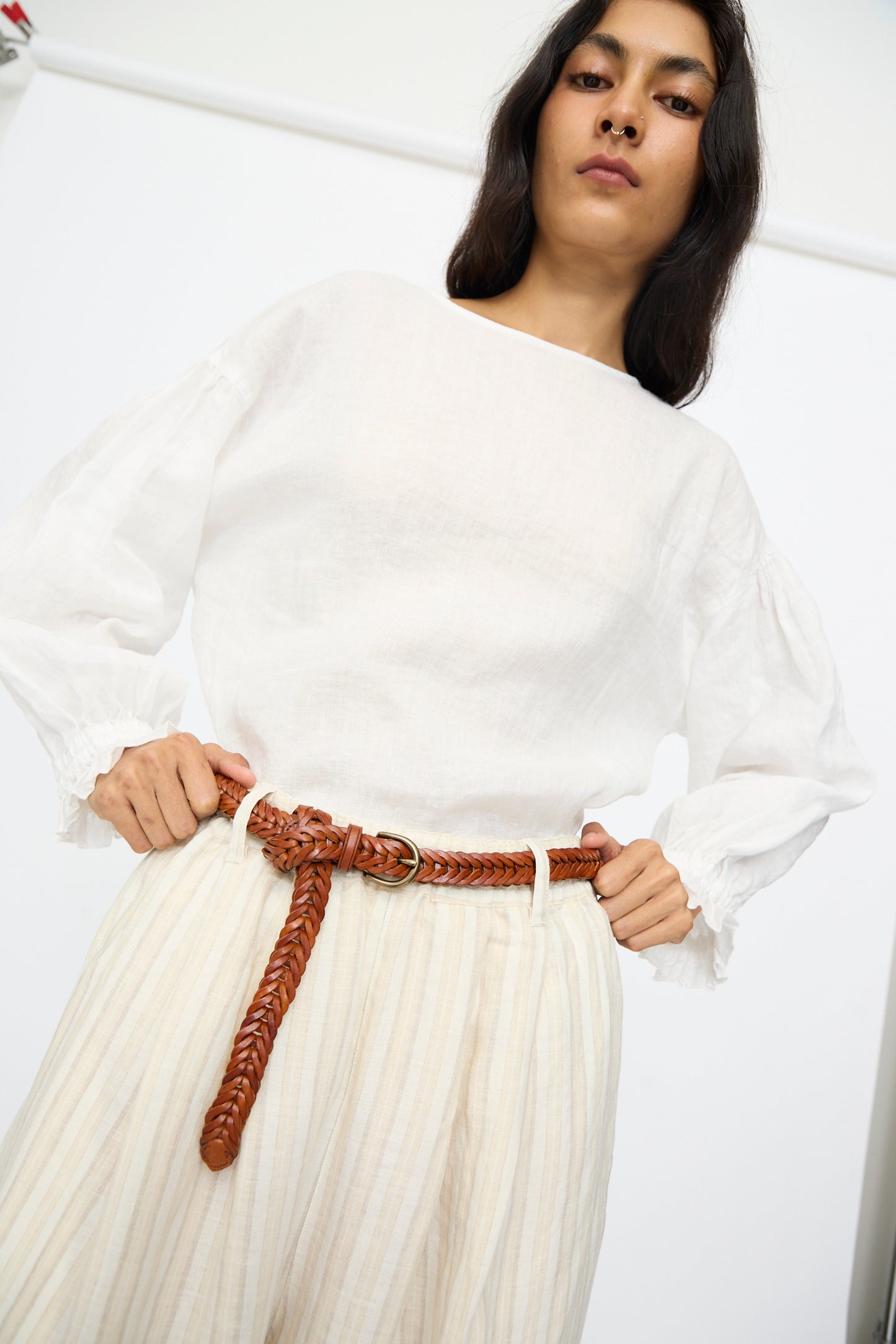 Wearing a sheer white blouse, light pleated pants, and the Brass Buckle Single Link Belt in Tan by Dragon Diffusion, this person stands hands on hips, looking down at the camera.