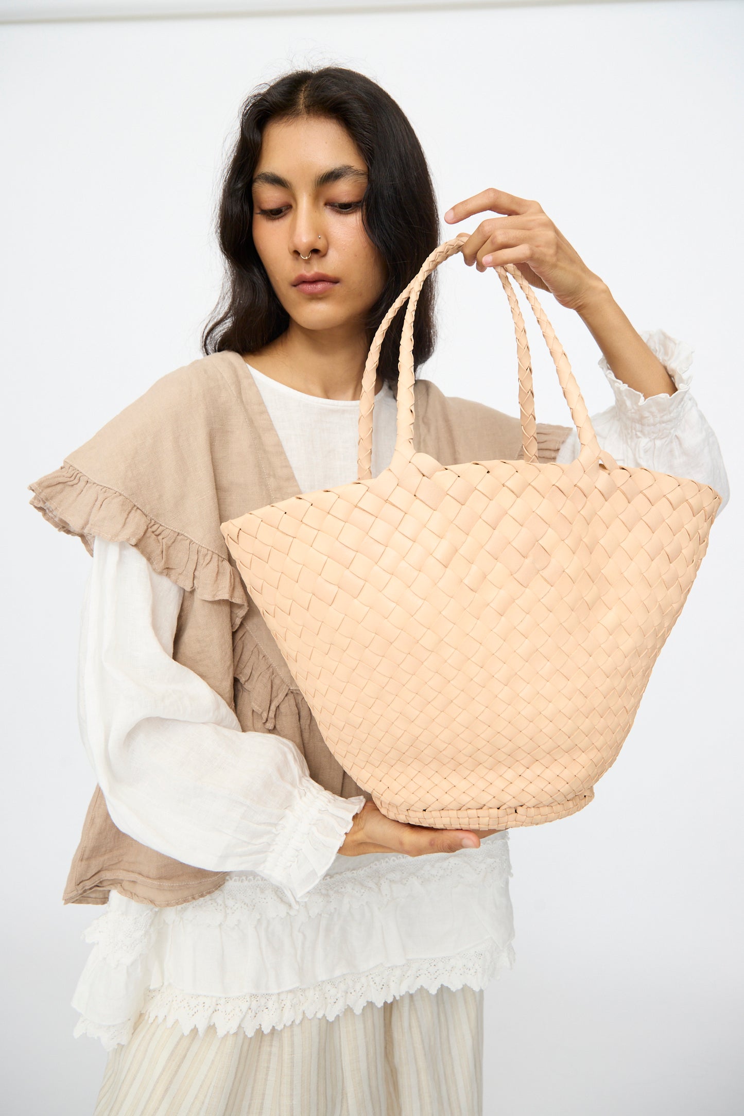 A woman in a white blouse and beige vest holds the Dragon Diffusion Egola in Natural, a large light-colored woven leather handbag made in India, against a plain background.