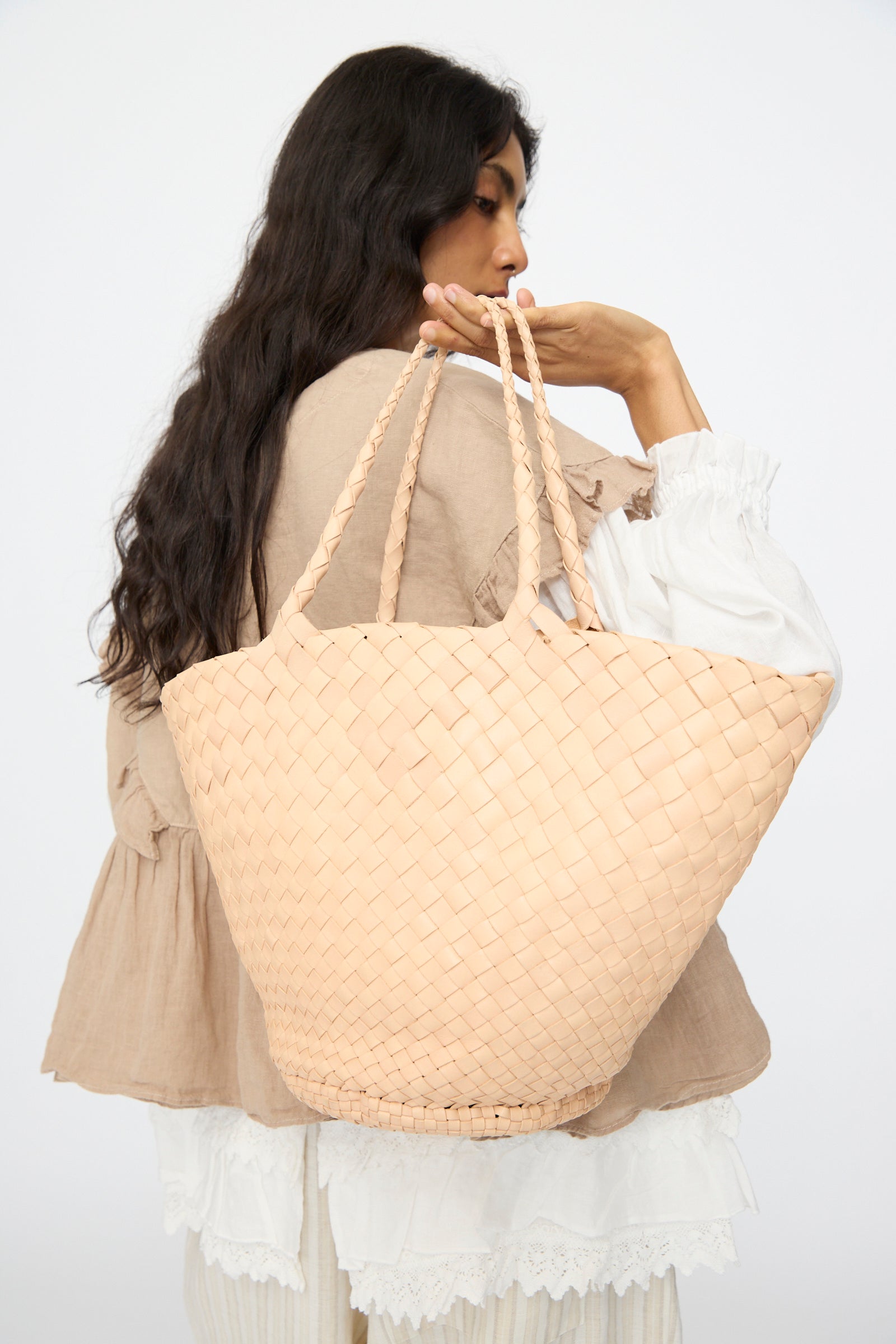 A woman with long dark hair carries the Dragon Diffusion Egola in Natural, a large woven leather handbag made in India, over her shoulder. She wears layered beige and white and is turned away from the camera.