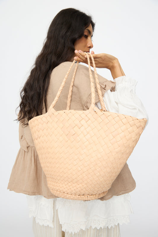 A woman with long dark hair carries the Dragon Diffusion Egola in Natural, a large woven leather handbag made in India, over her shoulder. She wears layered beige and white and is turned away from the camera.