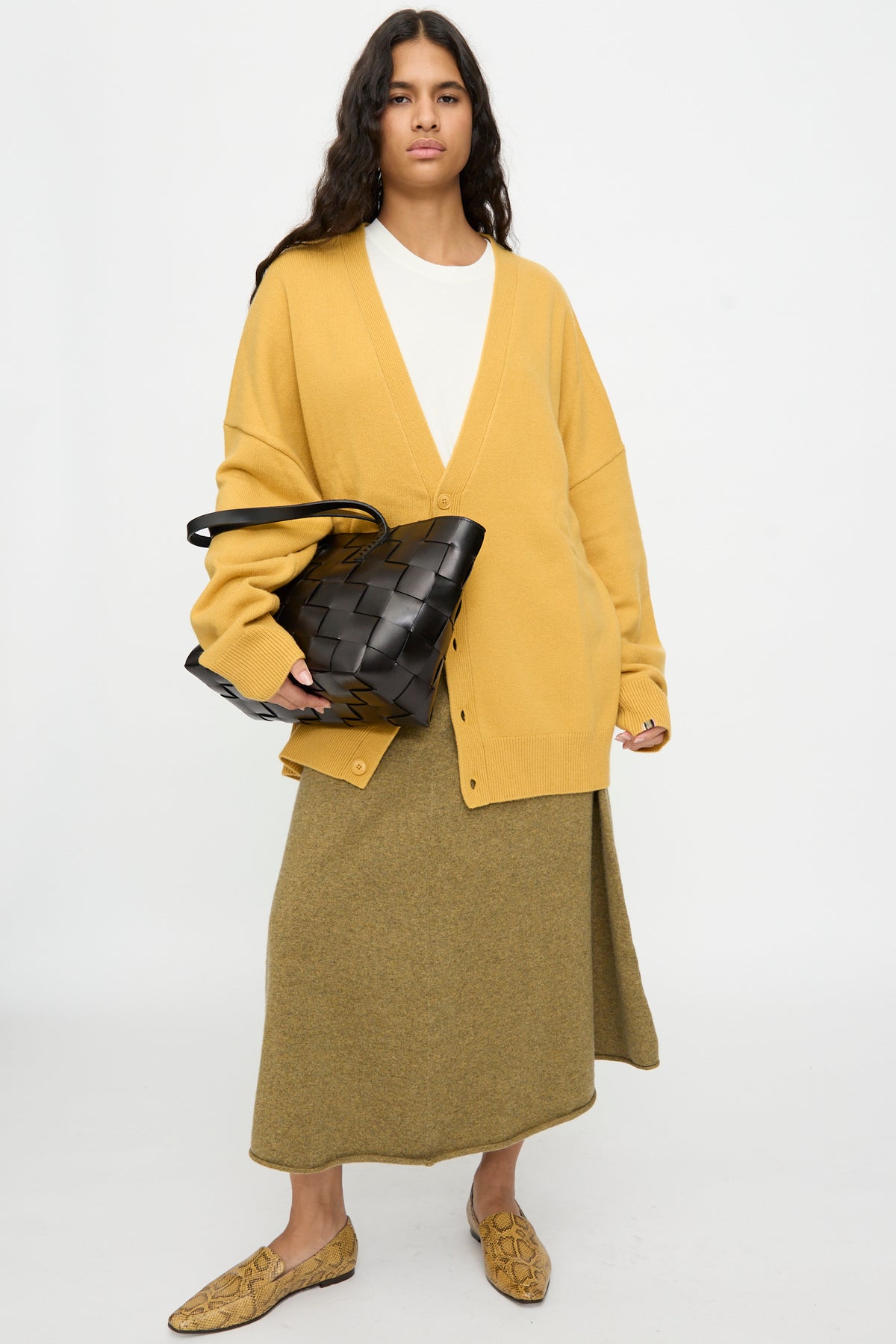 Person wearing a mustard yellow cardigan, olive skirt, white top, and snake-print loafers stands against a plain white background, holding the Dragon Diffusion Japan Tote in Black.