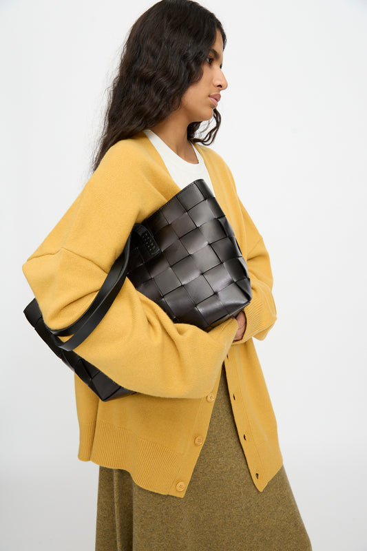 A woman in a yellow cardigan and olive skirt holds the Dragon Diffusion Japan Tote in Black—a large, basket-shaped bag crafted from cross-woven leather—against a plain white background.