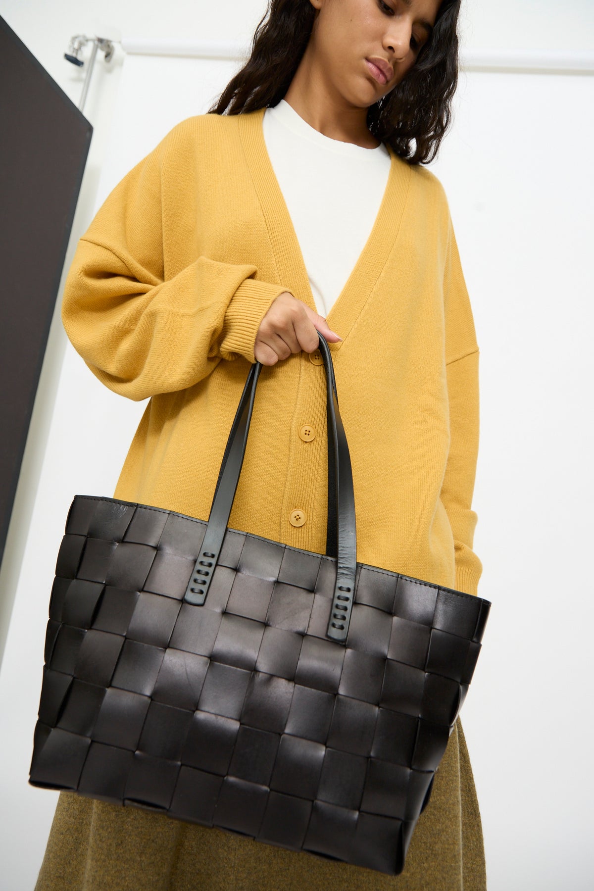 A woman in a mustard yellow cardigan holds the Dragon Diffusion Japan Tote in Black, a large cross-woven leather bag featuring two handles and a distinct basket shape, against a plain background.