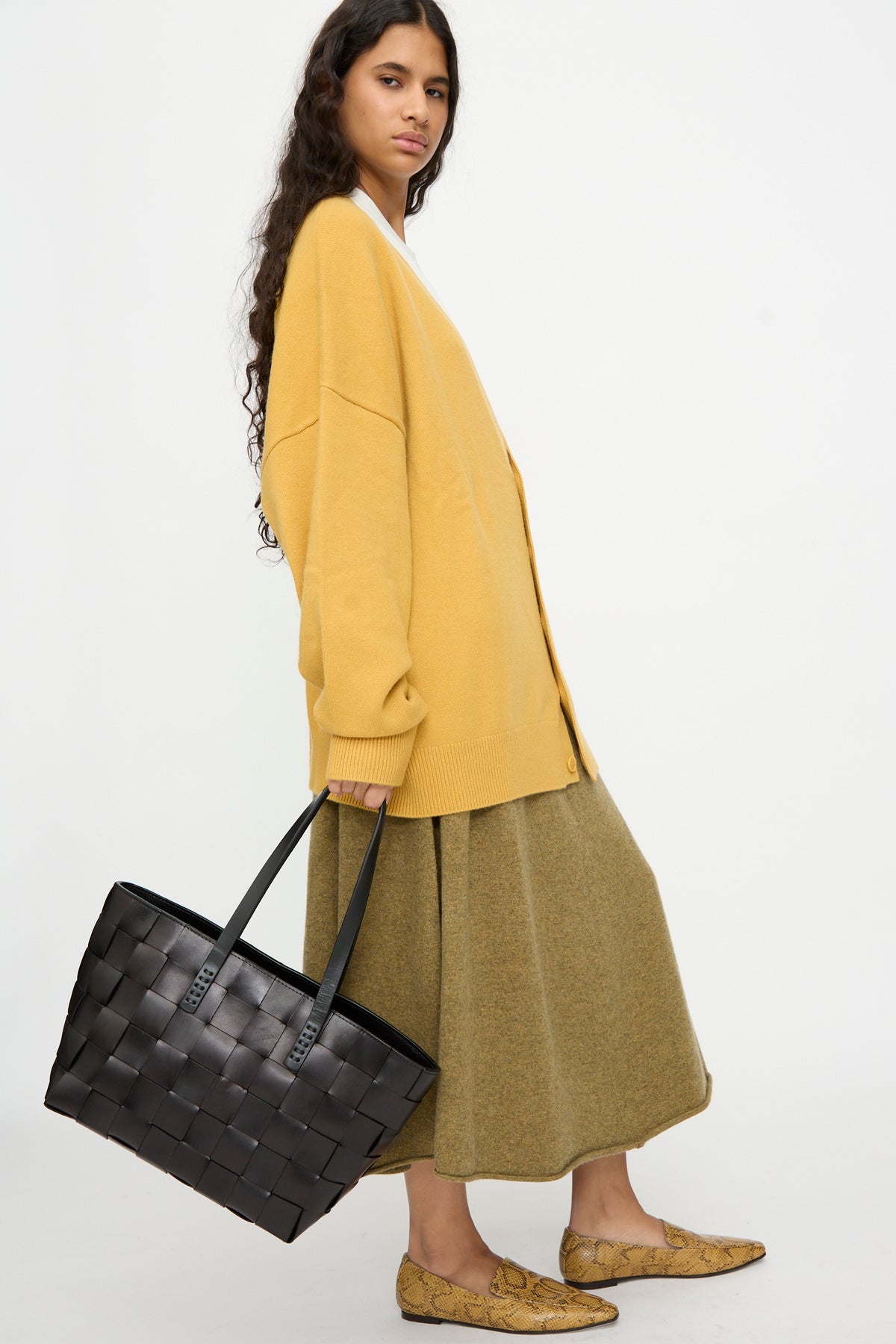 A person stands against a plain white background wearing an oversized yellow cardigan, olive skirt, and tan loafers, carrying the Dragon Diffusion Japan Tote in Black—a large, basket-shaped cross-woven leather bag.