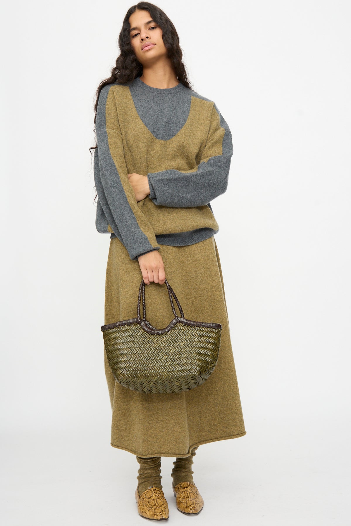 A person stands against a plain white background, wearing a gray and mustard colorblock sweater with a matching skirt and brown shoes, holding the Dragon Diffusion Nantucket Small Bi-Color bag in Dark Brown and Khaki.