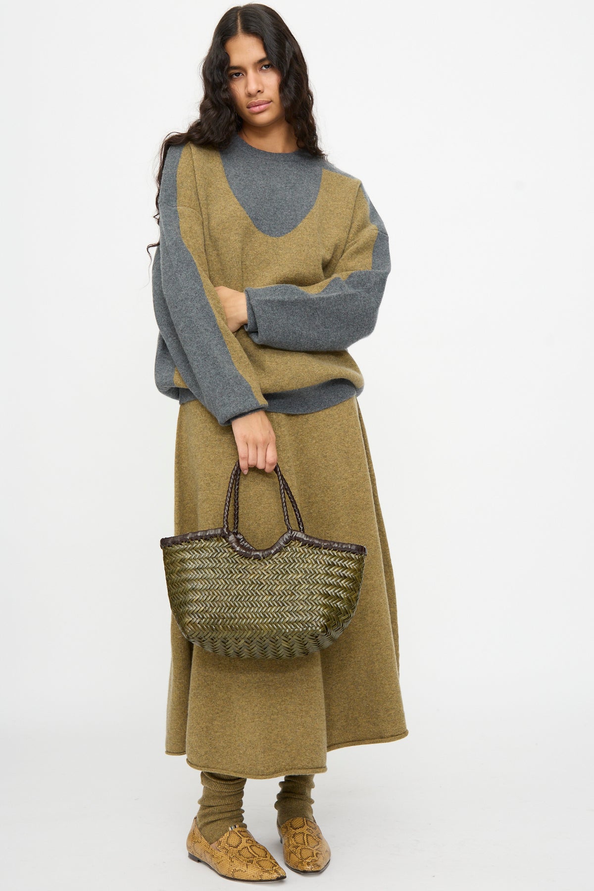 A person with long dark hair wears a gray and olive sweater, matching skirt, and shoes, holding the Dragon Diffusion Nantucket Small Bi-Color bag in Dark Brown and Khaki, standing against a plain white background.