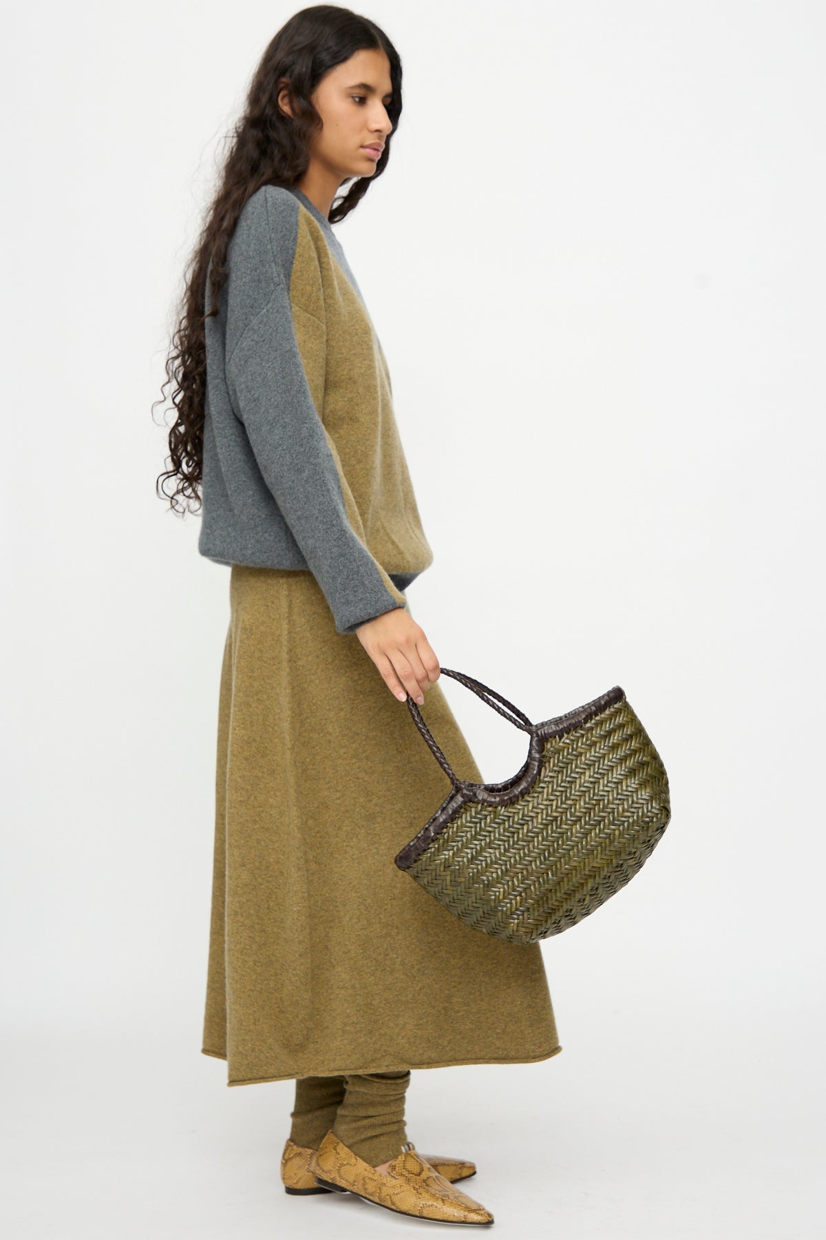 A person with long hair wears a grey and olive two-tone outfit and tan shoes, holding the Dragon Diffusion Nantucket Small Bi-Color bag in Dark Brown and Khaki while standing against a plain white background.