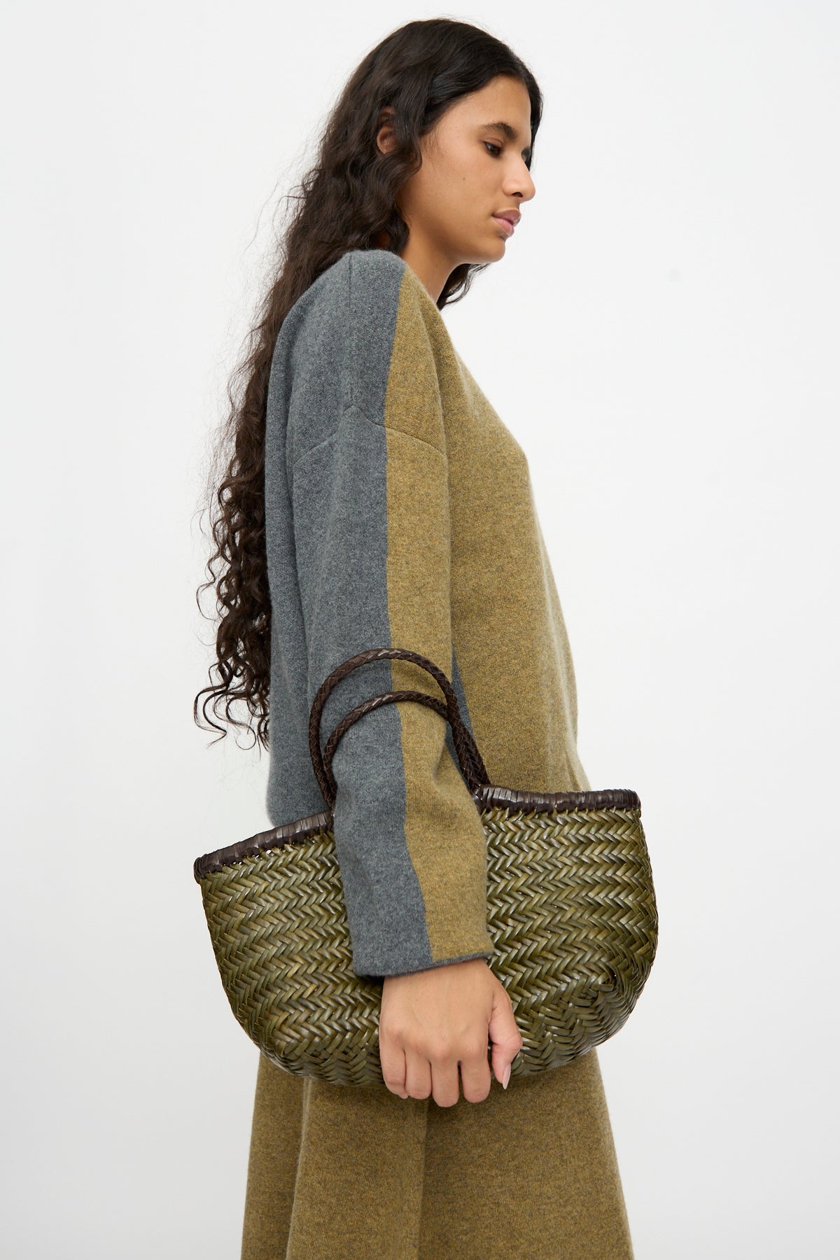 A woman with long, curly hair in a two-tone sweater dress holds the Dragon Diffusion Nantucket Small Bi-Color bag in Dark Brown and Khaki, featuring handwoven calf leather and dark handles, standing against a plain white background.