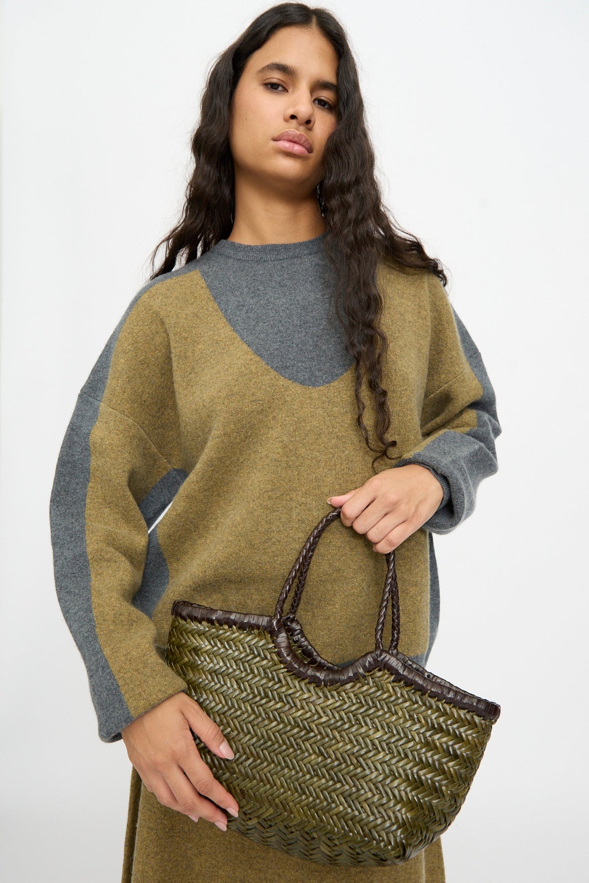 A person with long dark hair wears a brown and gray sweater dress and holds the Dragon Diffusion Nantucket Small Bi-Color bag in Dark Brown and Khaki, featuring a handwoven calf leather basket shape, against a plain background.