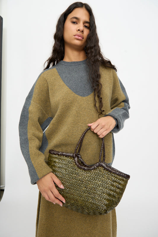 A person with long dark hair wears a brown and gray sweater dress and holds the Dragon Diffusion Nantucket Small Bi-Color bag in Dark Brown and Khaki, featuring a handwoven calf leather basket shape, against a plain background.