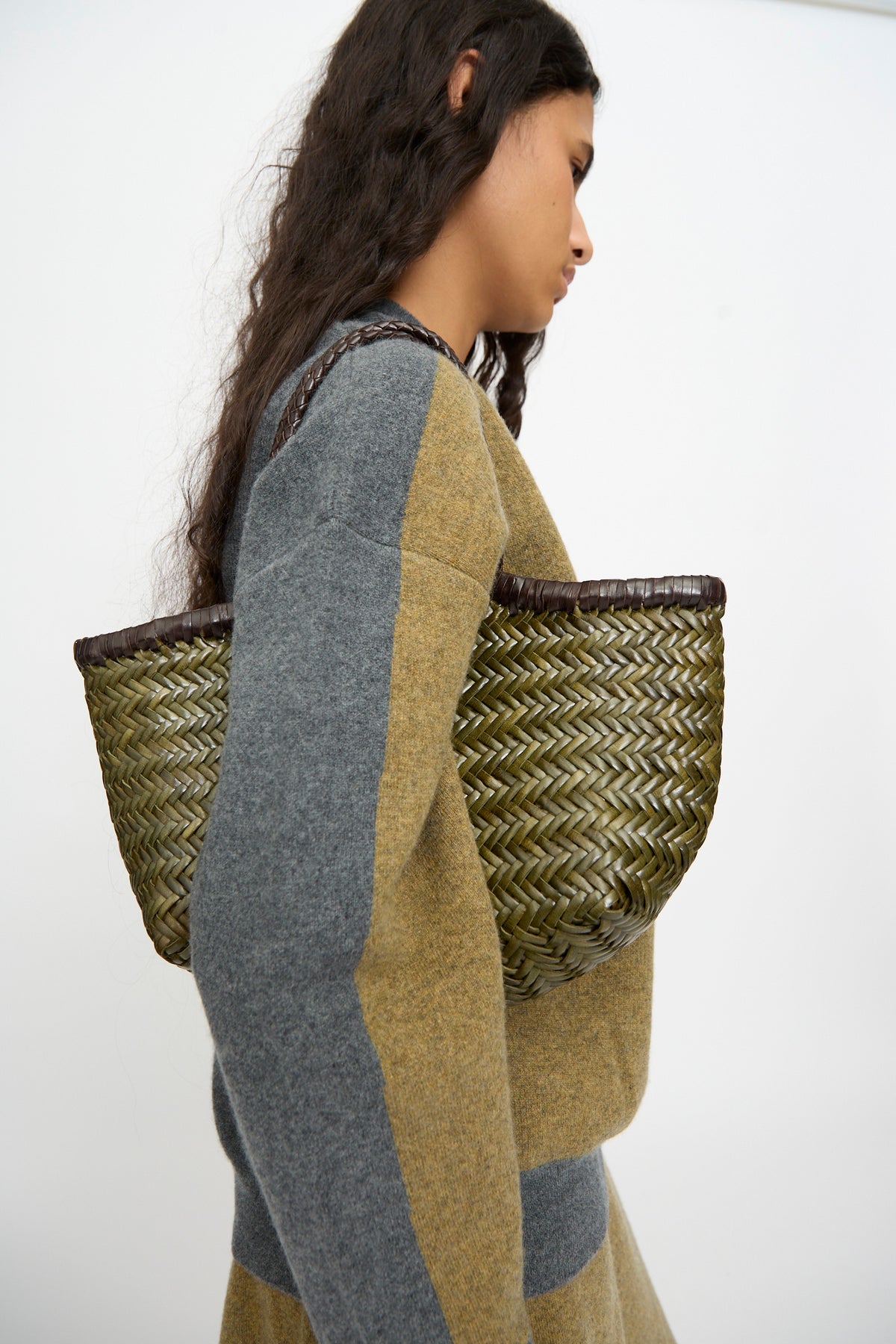 A woman with long, wavy hair wears a color-blocked sweater and skirt, carrying the Dragon Diffusion Nantucket Small Bi-Color bag in Dark Brown and Khaki—a handwoven calf leather basket—over her shoulder.