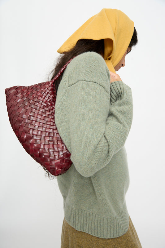 A person wearing a yellow headscarf, green sweater, and brown skirt carries the Dragon Diffusion Rosanna in Bordo handwoven calf leather bag over their shoulder, facing sideways against a plain background.