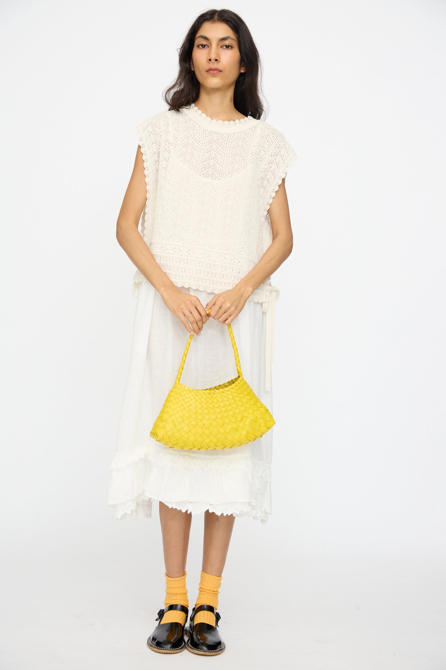 Person standing against a white background, wearing a white sleeveless knit top and skirt, yellow socks, black shoes, and holding the Dragon Diffusion Rosanna Bag in Yellow, handwoven from calf leather using basket weaving techniques.