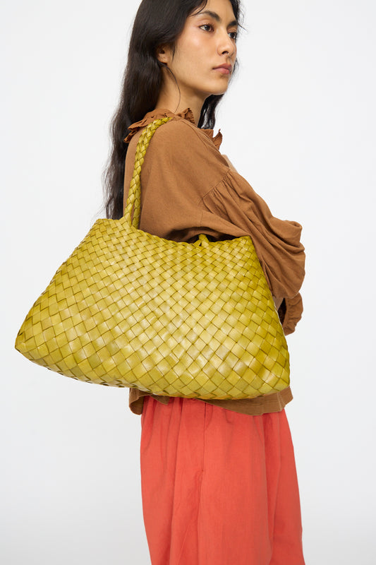 A person holds the Dragon Diffusion Santa Croce Big in Bamboo Green bag, featuring intricate handwoven basket design, over their shoulder while standing against a plain background.