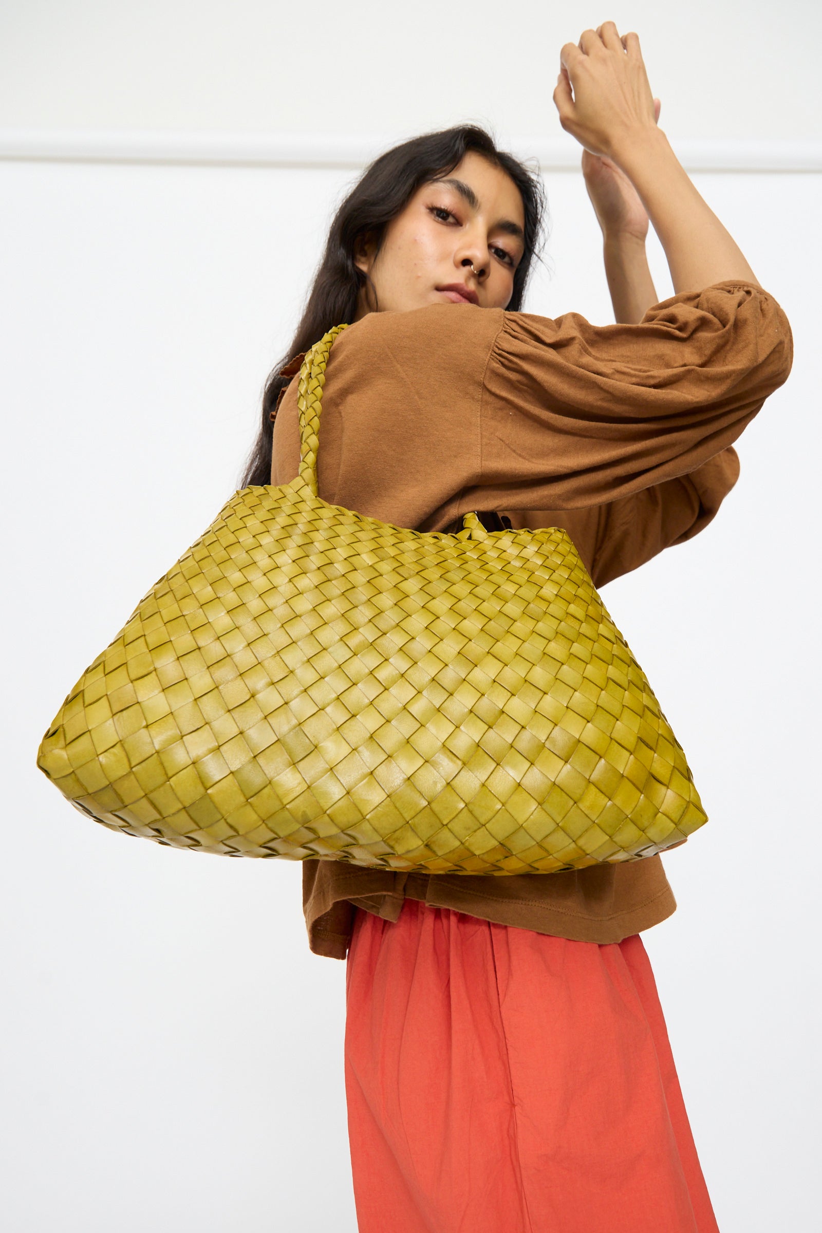 Wearing a brown top and orange skirt, a person poses with the Dragon Diffusion Santa Croce Big in Bamboo Green bag over their shoulder against a plain background.