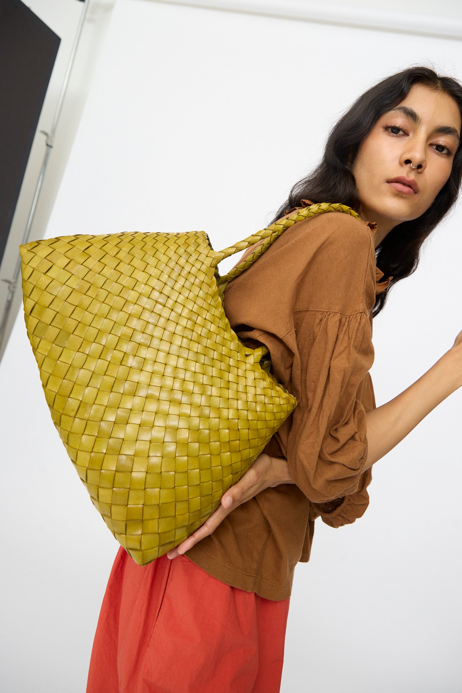 A woman in a brown top and red skirt poses with the Dragon Diffusion Santa Croce Big in Bamboo Green, a large handwoven bag with intricate basket weaving, slung over her shoulder against a plain background.