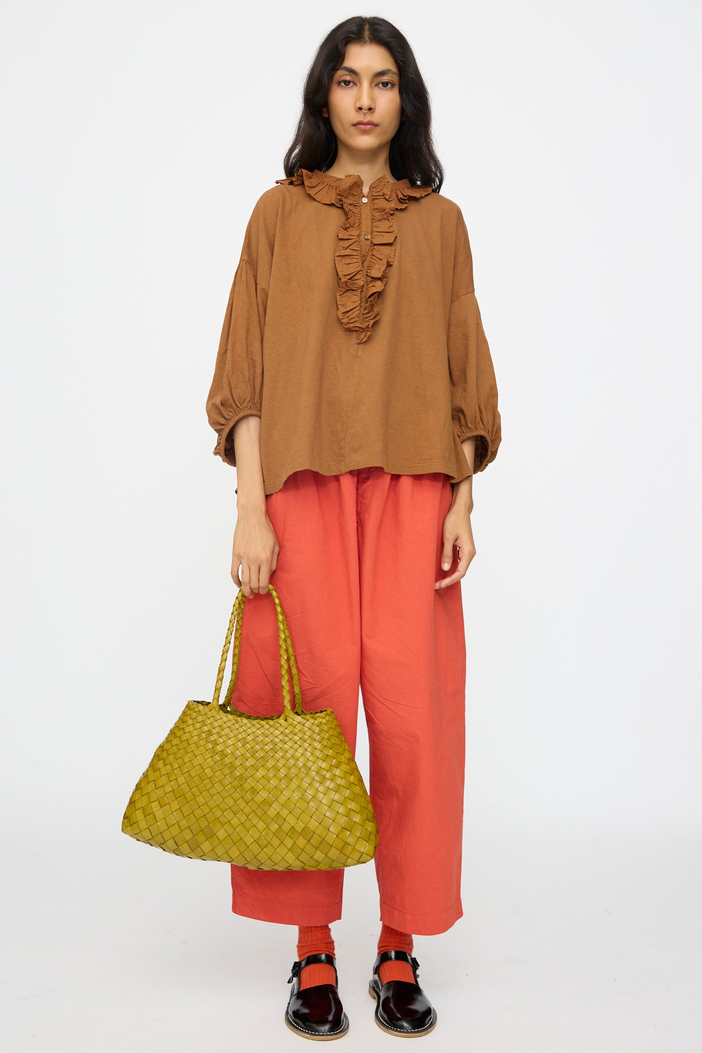 A woman stands against a plain background, wearing a brown ruffled blouse, loose red-orange pants, black shoes, and holding the Dragon Diffusion Santa Croce Big in Bamboo Green bag.