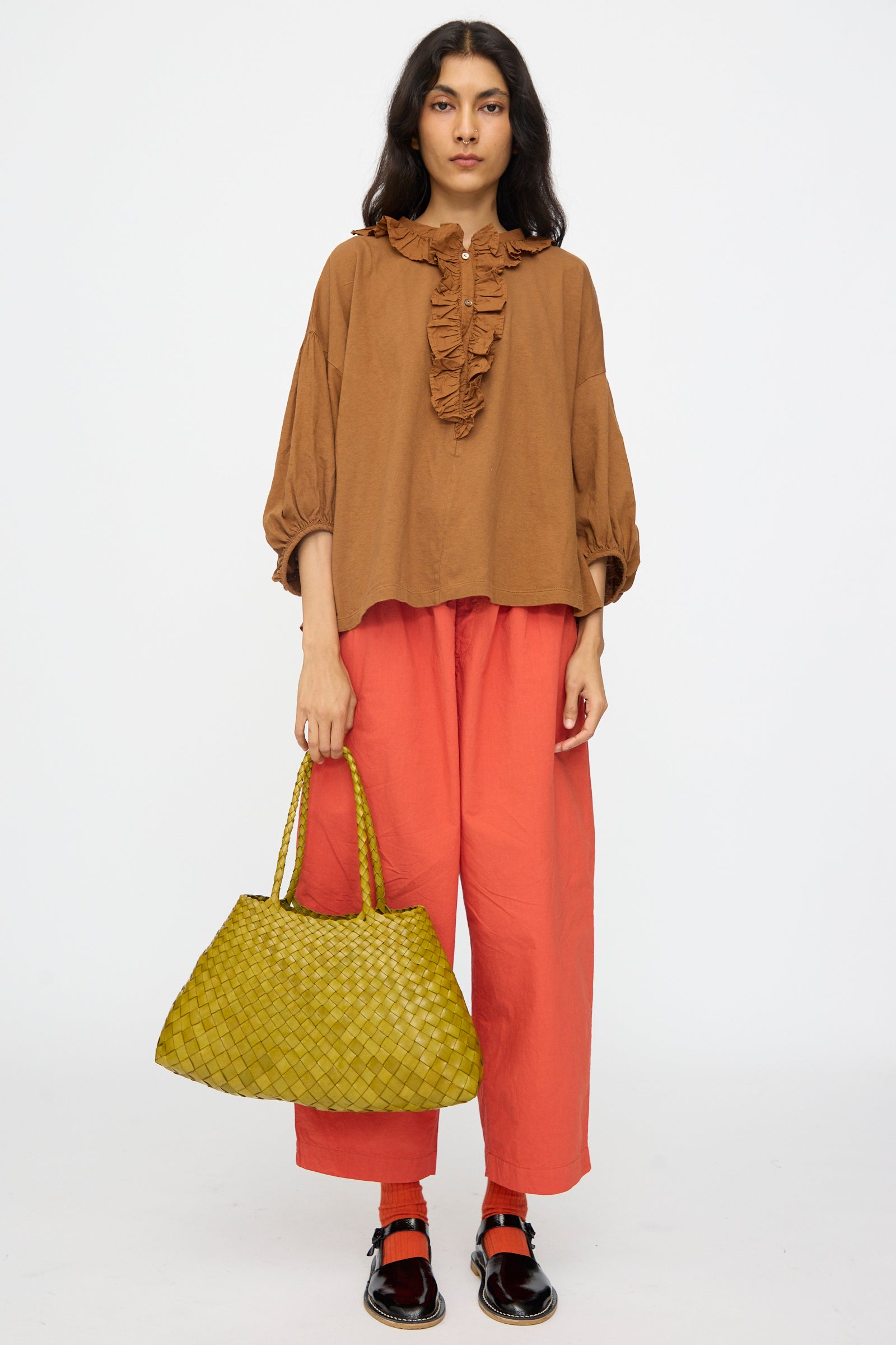 A woman stands against a plain background, wearing a brown ruffled blouse, loose red-orange pants, black shoes, and holding the Dragon Diffusion Santa Croce Big in Bamboo Green bag.
