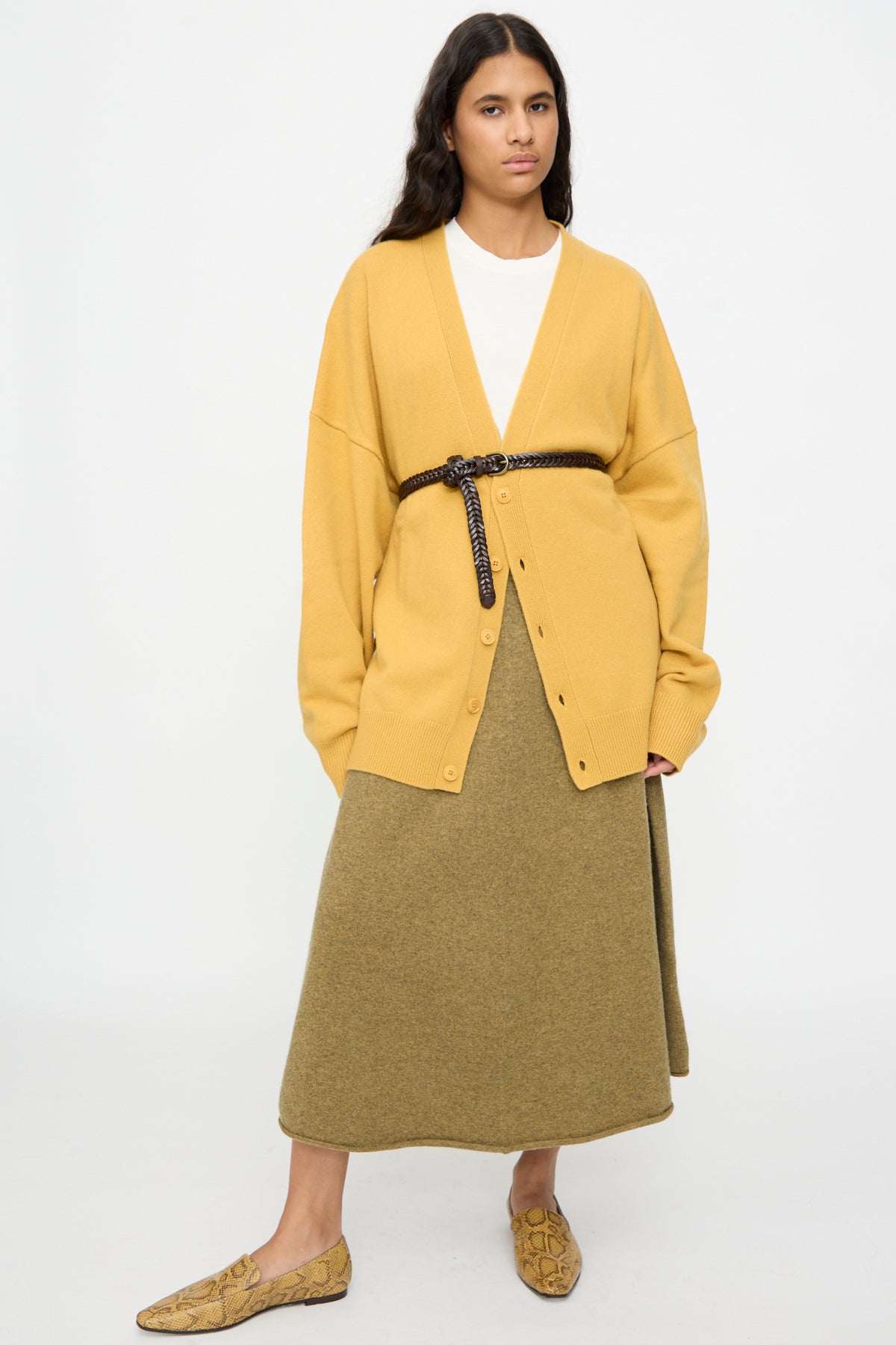 A person wearing a yellow cardigan cinched with the Dragon Diffusion Single Link Belt in Brown, a light olive skirt, tan snake-print loafers, and a white shirt stands against a plain white background.