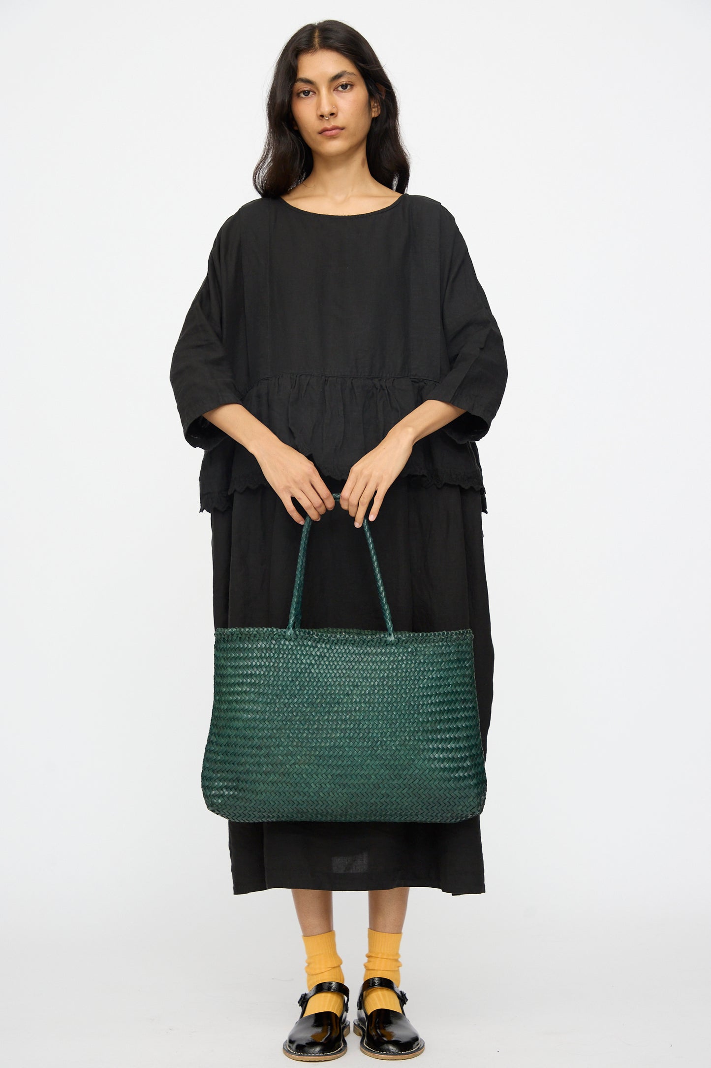 A woman in a loose black dress faces forward, holding the Dragon Diffusion Sophie Large in Forest handwoven tote with braided handles. She pairs yellow socks with black sandals against a plain white background.