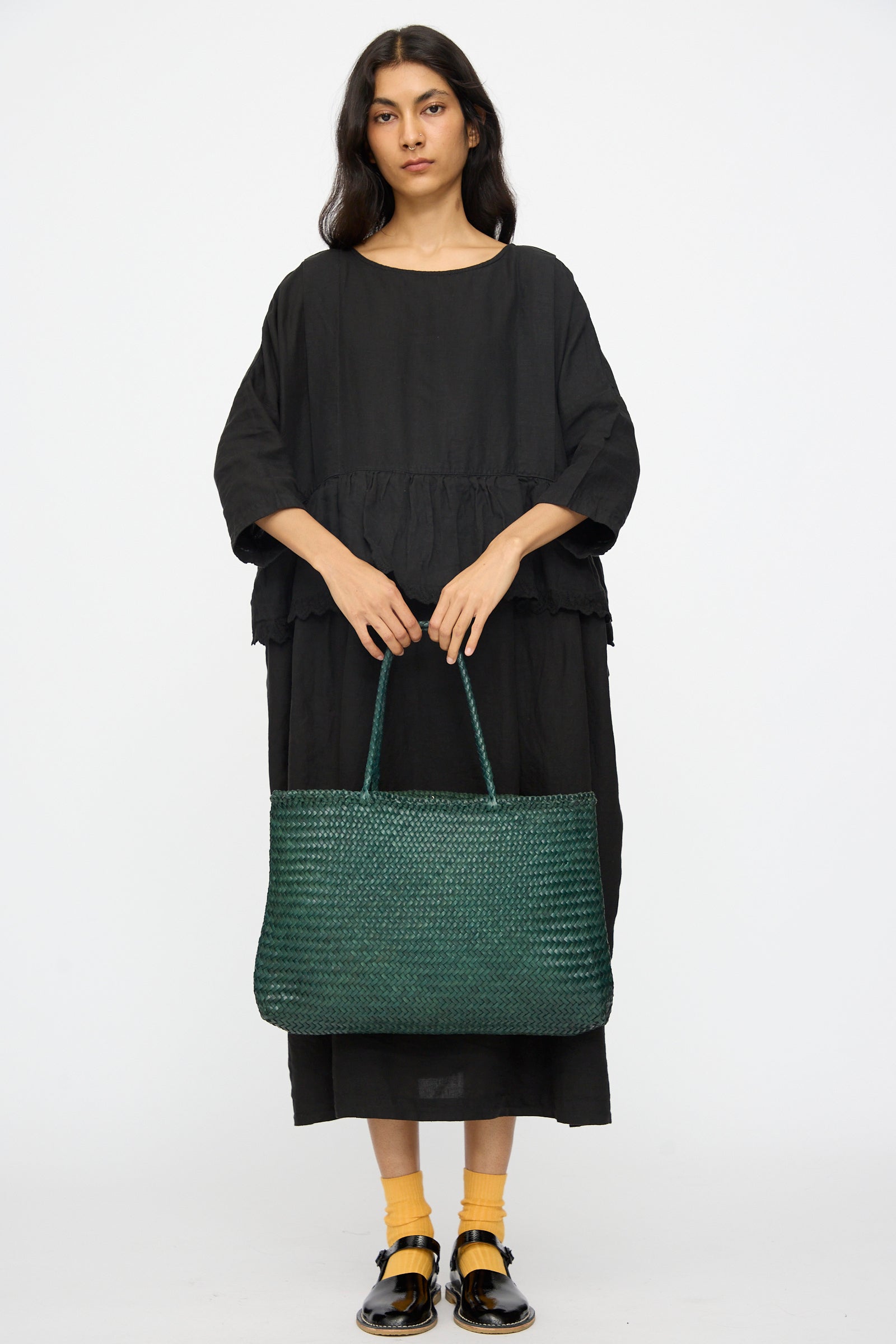 A woman in a loose black dress faces forward, holding the Dragon Diffusion Sophie Large in Forest handwoven tote with braided handles. She pairs yellow socks with black sandals against a plain white background.