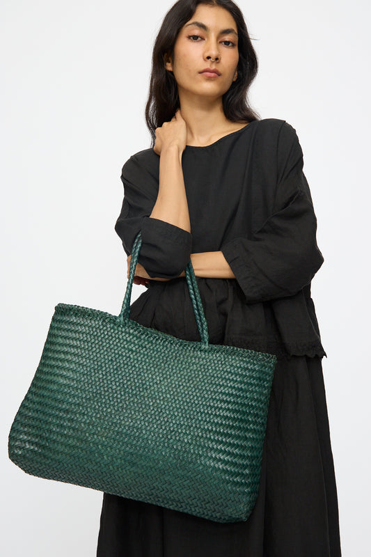 A person in a loose black dress holds the Dragon Diffusion Sophie Large tote in Forest, featuring braided handles, against a plain white background.