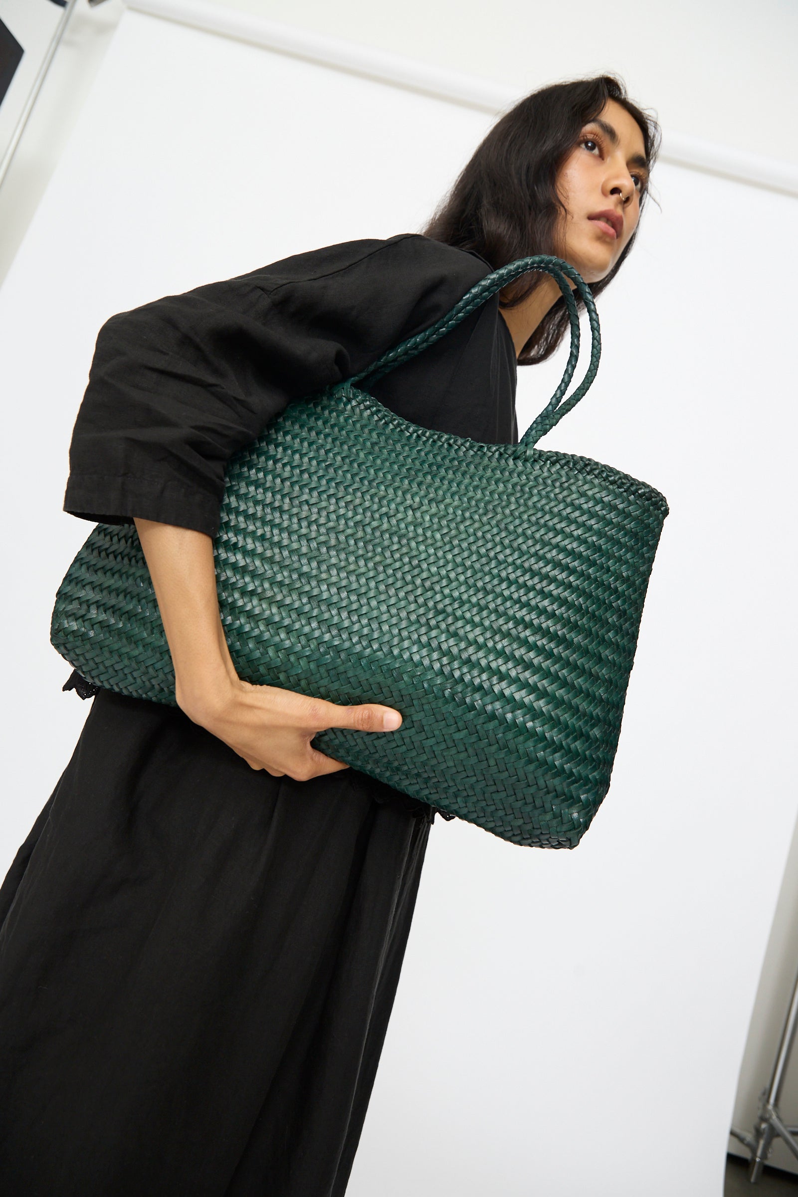 A woman in a black dress holds the Dragon Diffusion "Sophie Large in Forest" handwoven tote with braided handles, standing against a plain white background.