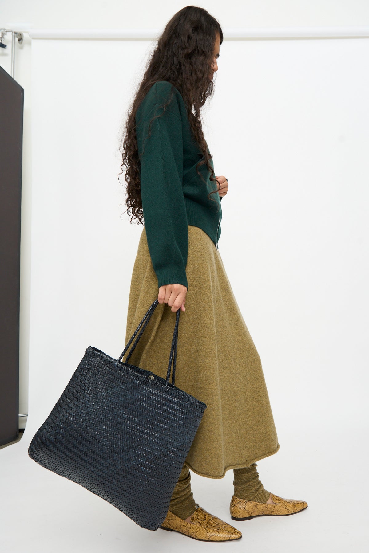 Person with long hair wears a green sweater, olive skirt, textured leggings, snakeskin boots, and carries the Dragon Diffusion Sophie Large in Marine handwoven calf leather tote bag against a plain white background.