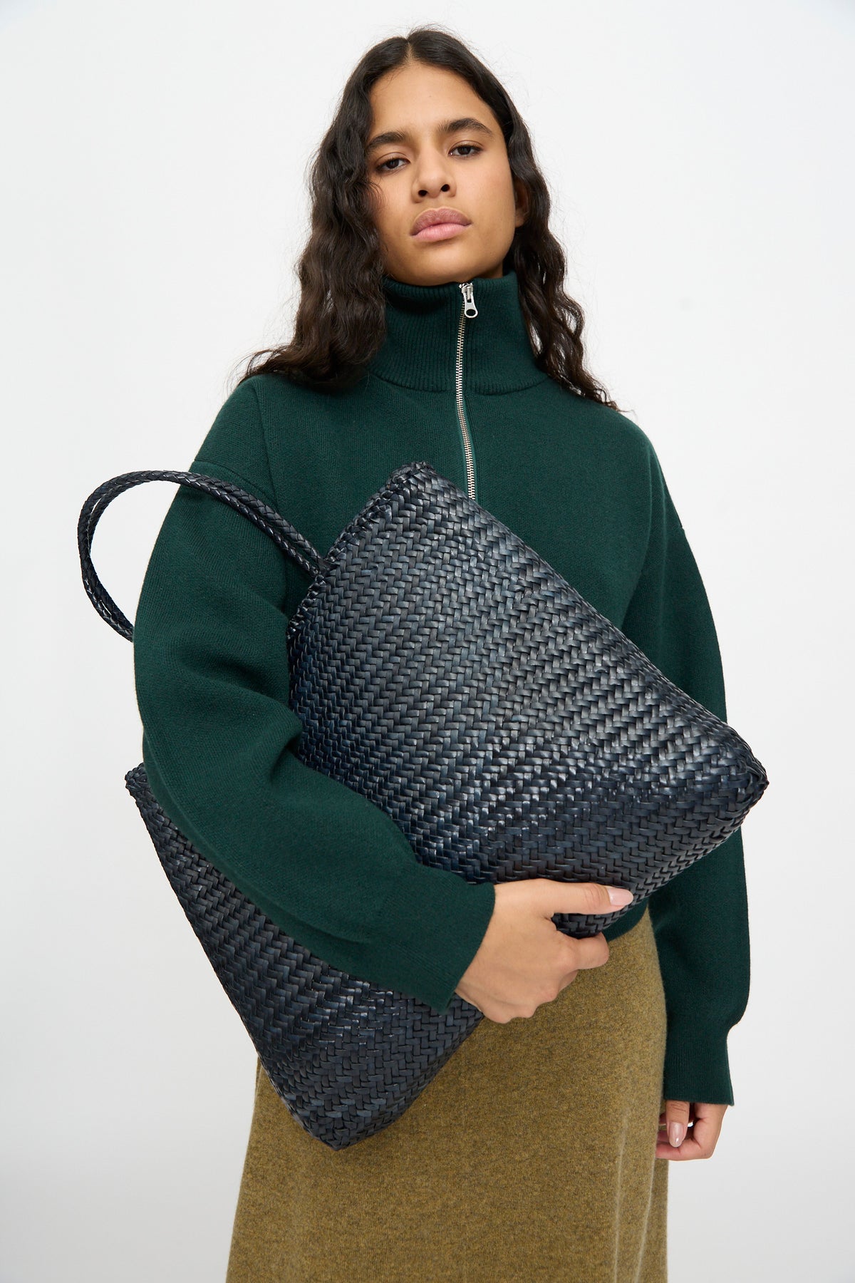 A woman in a dark green zip-up sweater and brown skirt holds the Dragon Diffusion Sophie Large tote in Marine, a handwoven calf leather bag, against a plain white background.