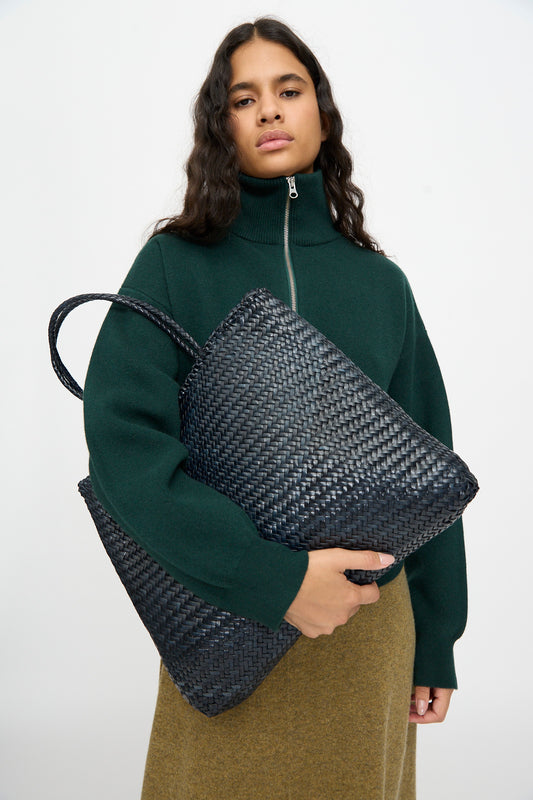 A woman in a dark green zip-up sweater and brown skirt holds the Dragon Diffusion Sophie Large tote in Marine, a handwoven calf leather bag, against a plain white background.