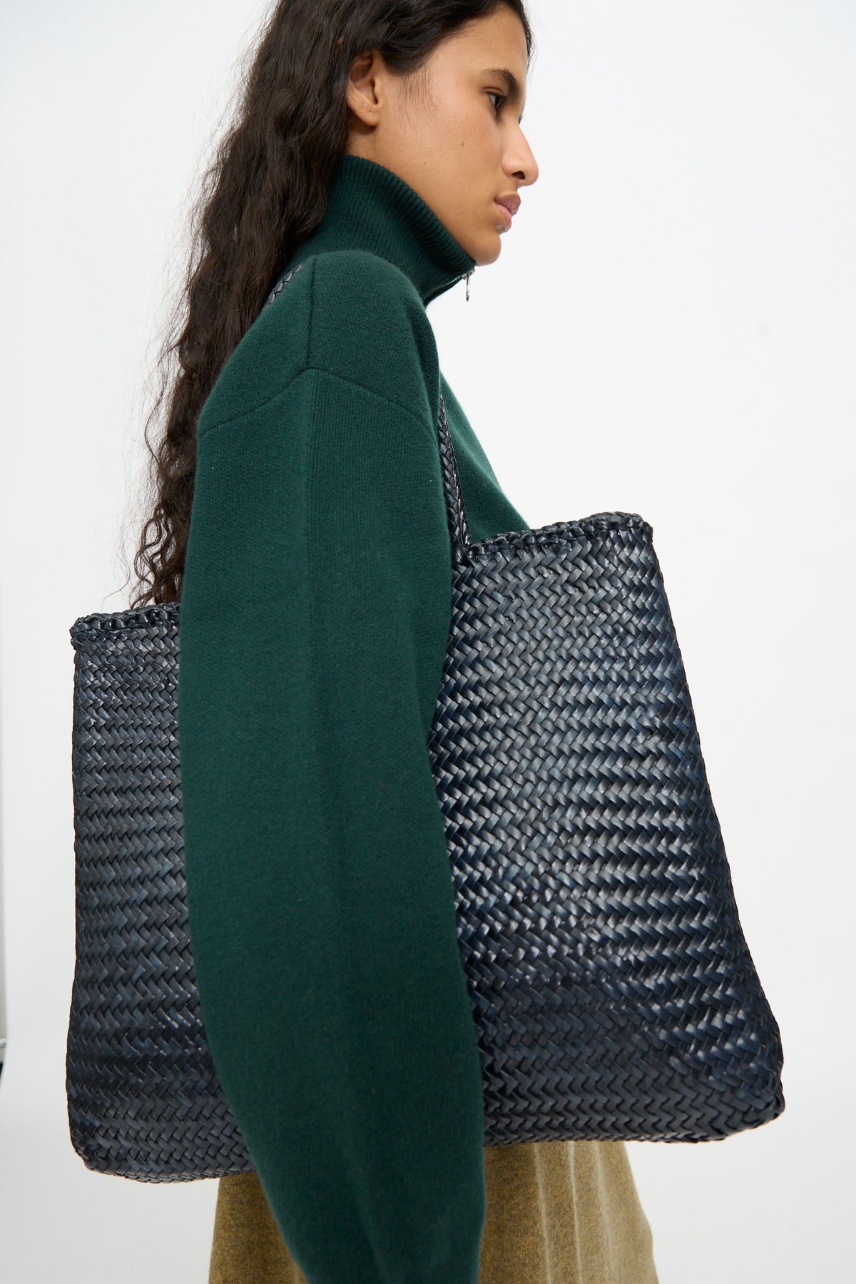 A person wearing a green turtleneck sweater carries the Dragon Diffusion Sophie Large in Marine, a handwoven calf leather tote bag, over their shoulder.