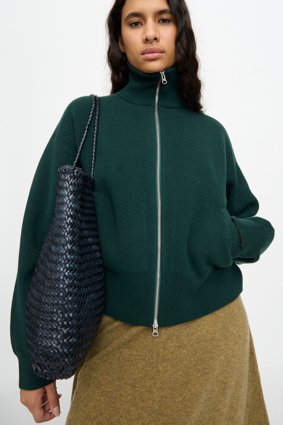 A woman stands against a plain background, wearing a dark green zip-up sweater and tan skirt, and carrying the Dragon Diffusion Sophie Large in Marine, a handwoven calf leather tote bag.