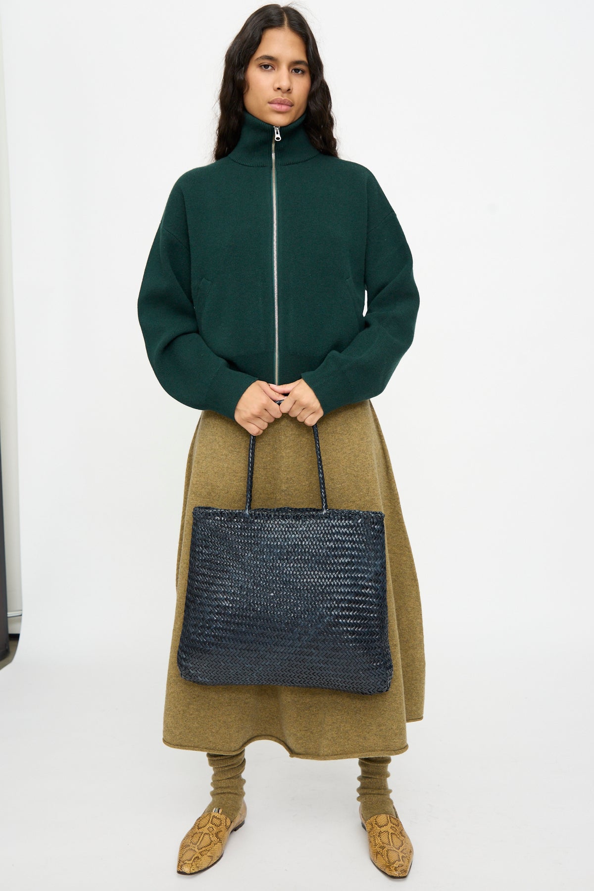 A woman stands against a plain white background wearing a dark green zip-up jacket, tan midi skirt, and tan patterned shoes, while holding the Dragon Diffusion Sophie Large in Marine—a handwoven calf leather tote bag.
