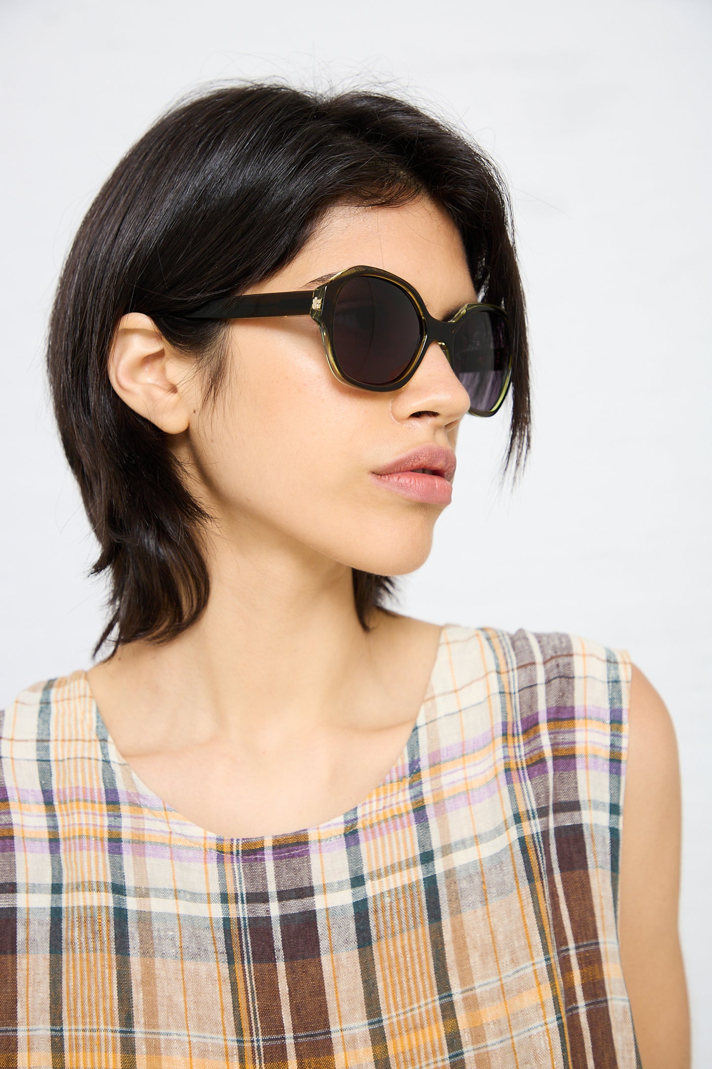 Woman with short dark hair wearing Eva Masaki's 001 Sunglasses in Greens and a plaid sleeveless top, looking to the side against a plain background. The sunglasses offer 100% UV protection, ensuring both style and safety.