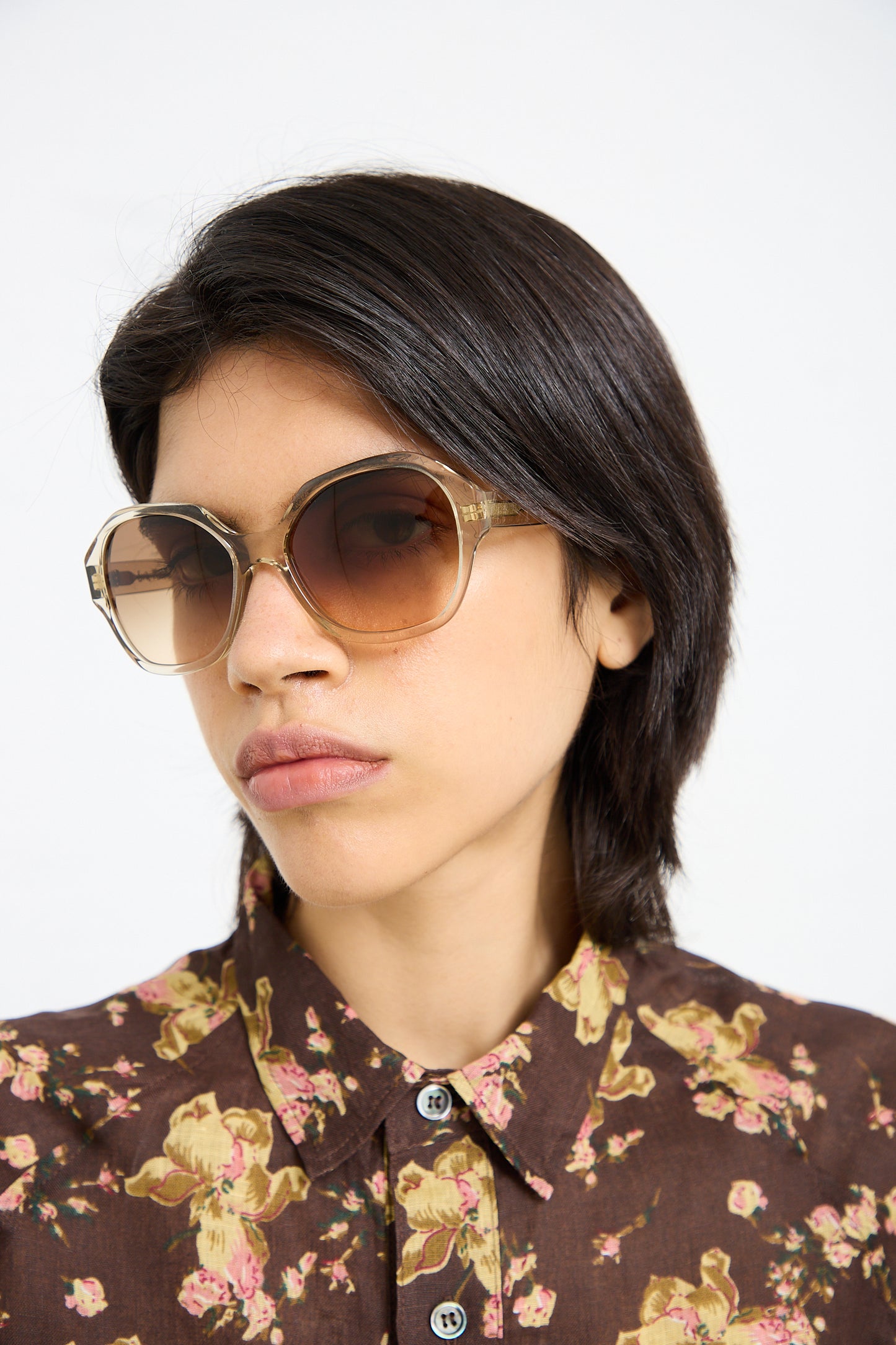 Person with short dark hair wearing large 001 Sunglasses in Toast by Eva Masaki and a floral-patterned shirt.