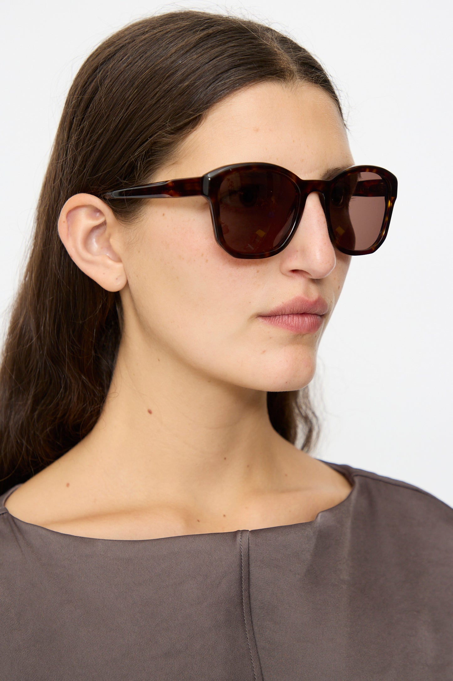 Woman with long brown hair wears Eva Masaki BQE Sunglasses in Finch (oversized Havana tortoise shell) and a gray top, gazing slightly right against a plain background.