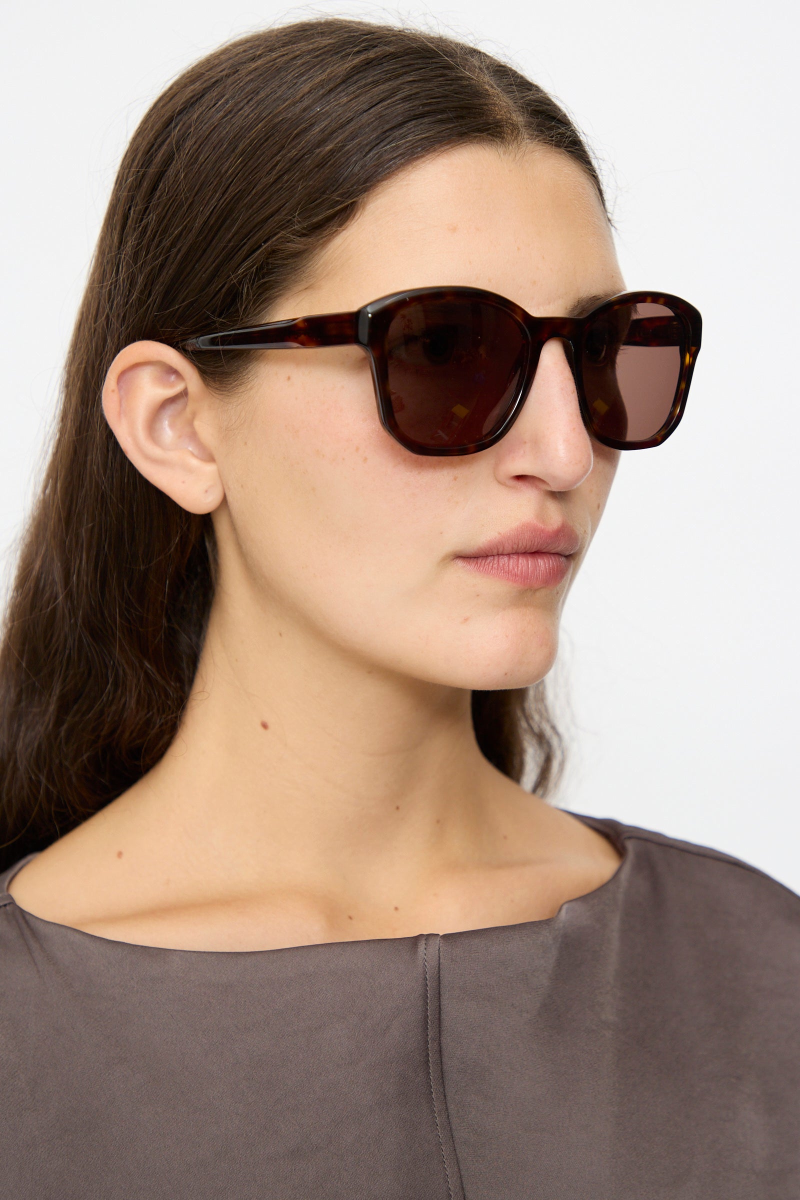 Woman with long brown hair wears Eva Masaki BQE Sunglasses in Finch (oversized Havana tortoise shell) and a gray top, gazing slightly right against a plain background.