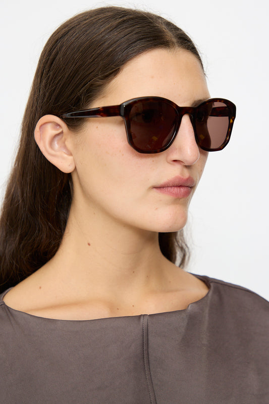 Woman with long brown hair wears Eva Masaki BQE Sunglasses in Finch (oversized Havana tortoise shell) and a gray top, gazing slightly right against a plain background.