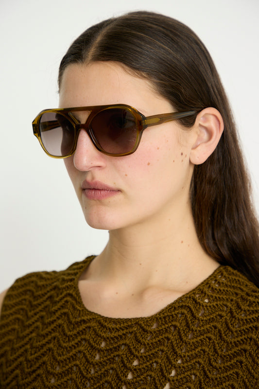 A person with long dark hair dons oversized Chiyo Aviator Sunglasses in Avocado by Eva Masaki, paired with a stylish brown knit top against a plain background.