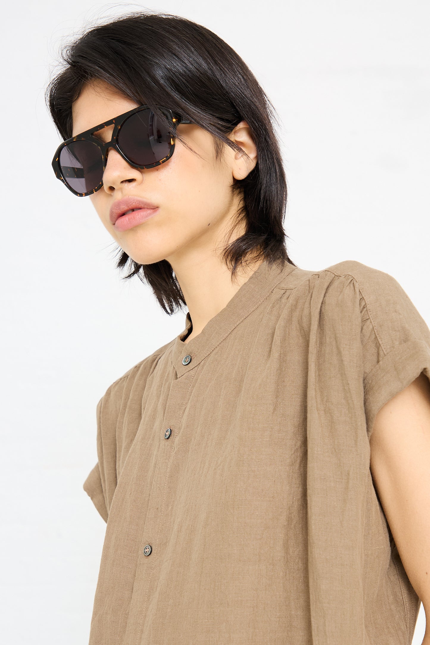 Wearing the chic Chiyo Aviator Sunglasses in Gotham by Eva Masaki, paired with a brown button-up shirt, a person stands confidently against a plain background.