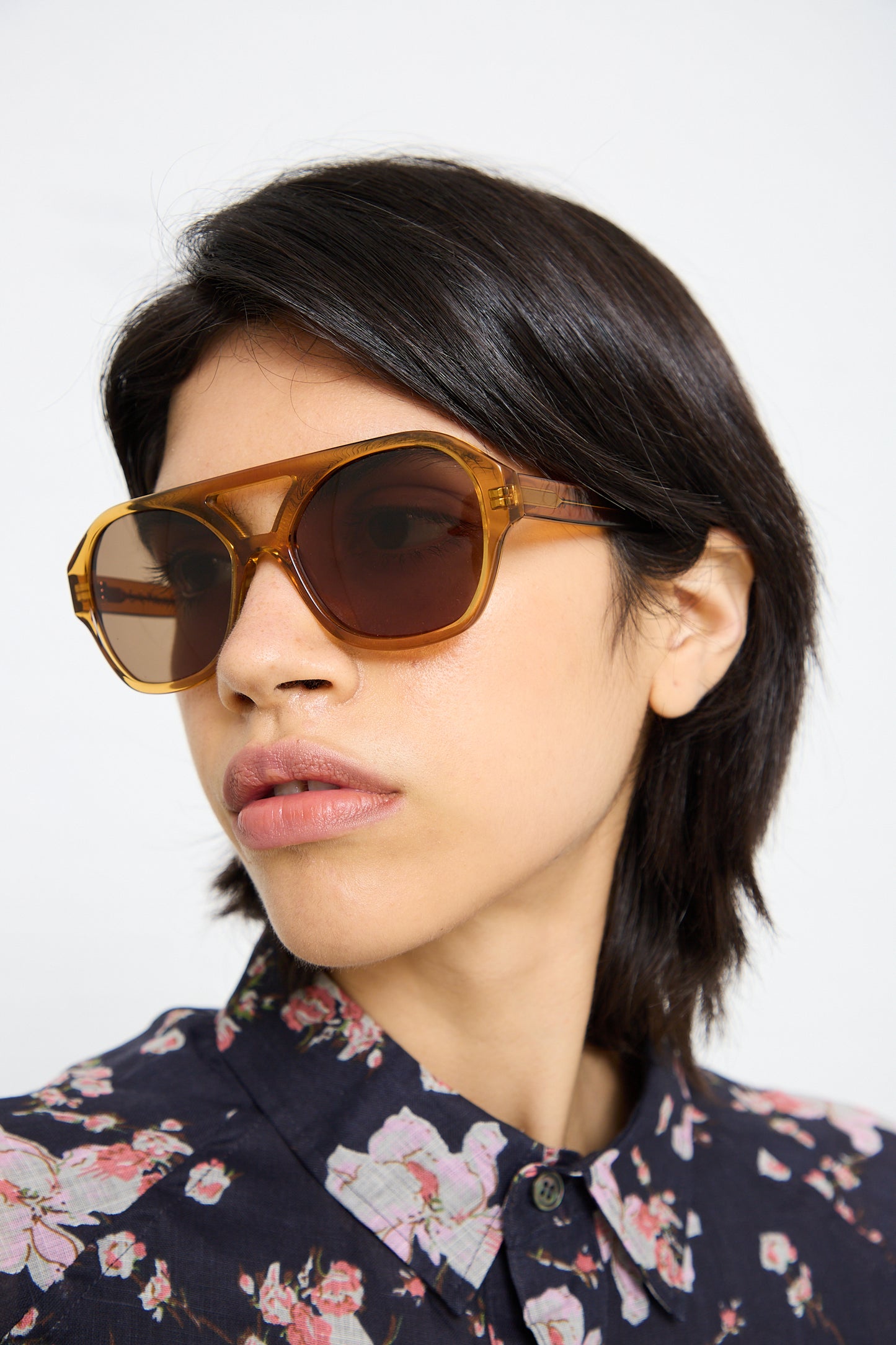 A person with short dark hair wearing large Eva Masaki Chiyo Aviator Sunglasses in Honey and a floral-patterned shirt looks to the side.