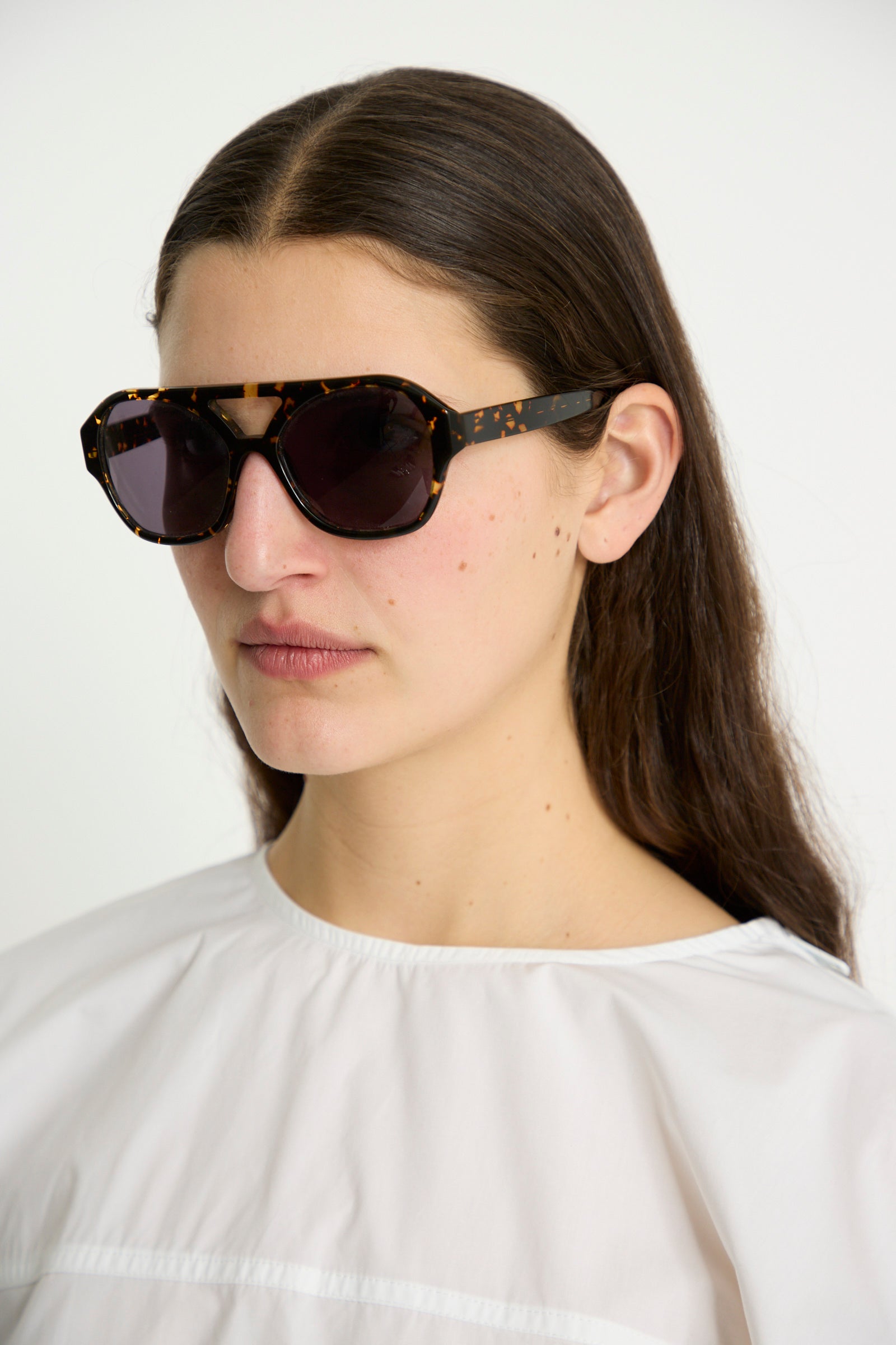 A person in a white shirt wearing Eva Masaki's Chiyo Aviator Sunglasses in Gotham, crafted from premium Italian acetate, gazes straight ahead against a plain background.