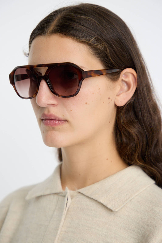 A woman with long brown hair is seen wearing large Chiyo Sunglasses in Good Luck by Eva Masaki, featuring hexagonal aviator frames crafted from Italian acetate and offering 100% UV protection. She pairs these distinctive sunglasses with a beige collared top, posing slightly to the left against a plain background.