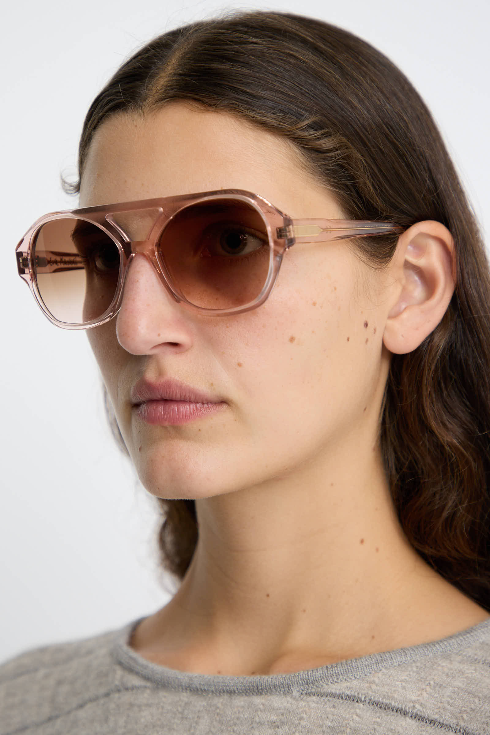 A person with long brown hair is wearing the Chiyo Aviators in Heart by Eva Masaki, featuring large, transparent pink frames crafted from Italian acetate, paired with a gray top, looking to the side.