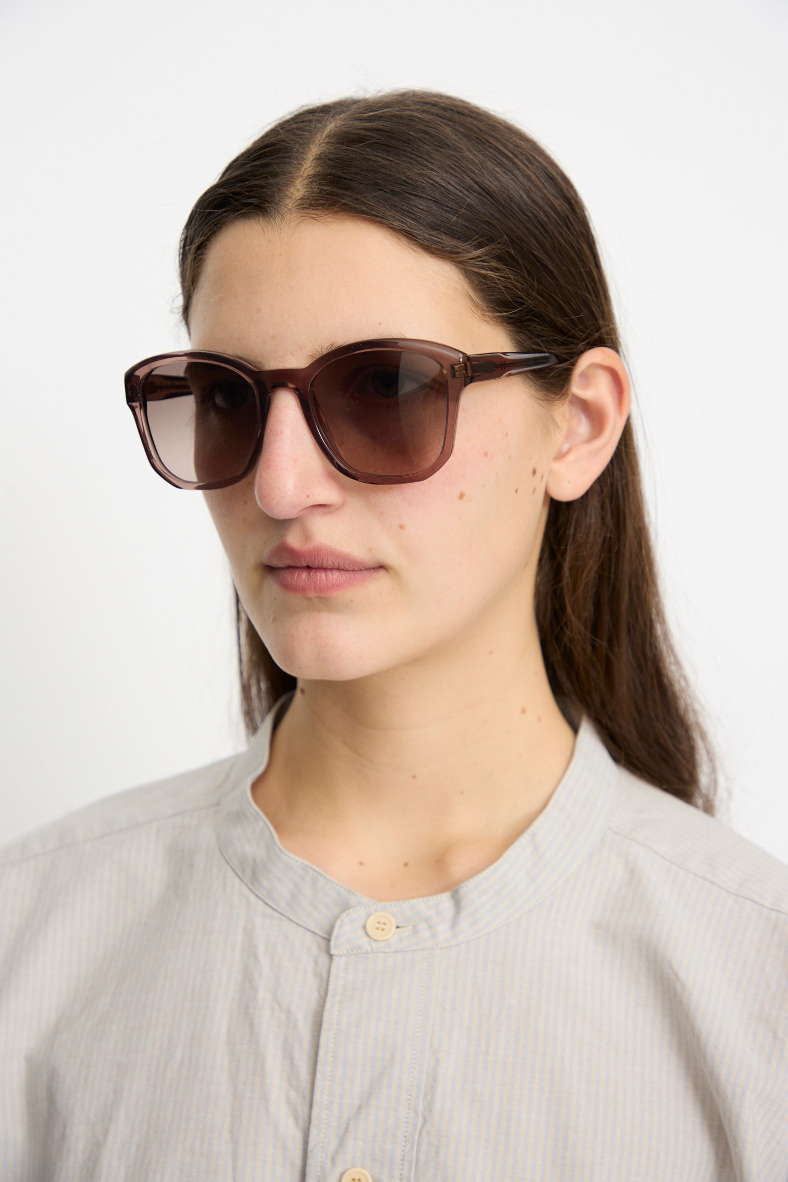 A person with long dark hair wears Eva Masaki BQE Sunglasses in Bambi with Zeiss lenses and a light-colored buttoned shirt against a plain background.