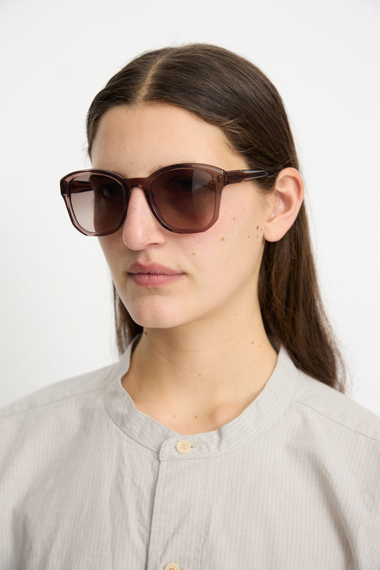 A person with long dark hair wears Eva Masaki BQE Sunglasses in Bambi with Zeiss lenses and a light-colored buttoned shirt against a plain background.