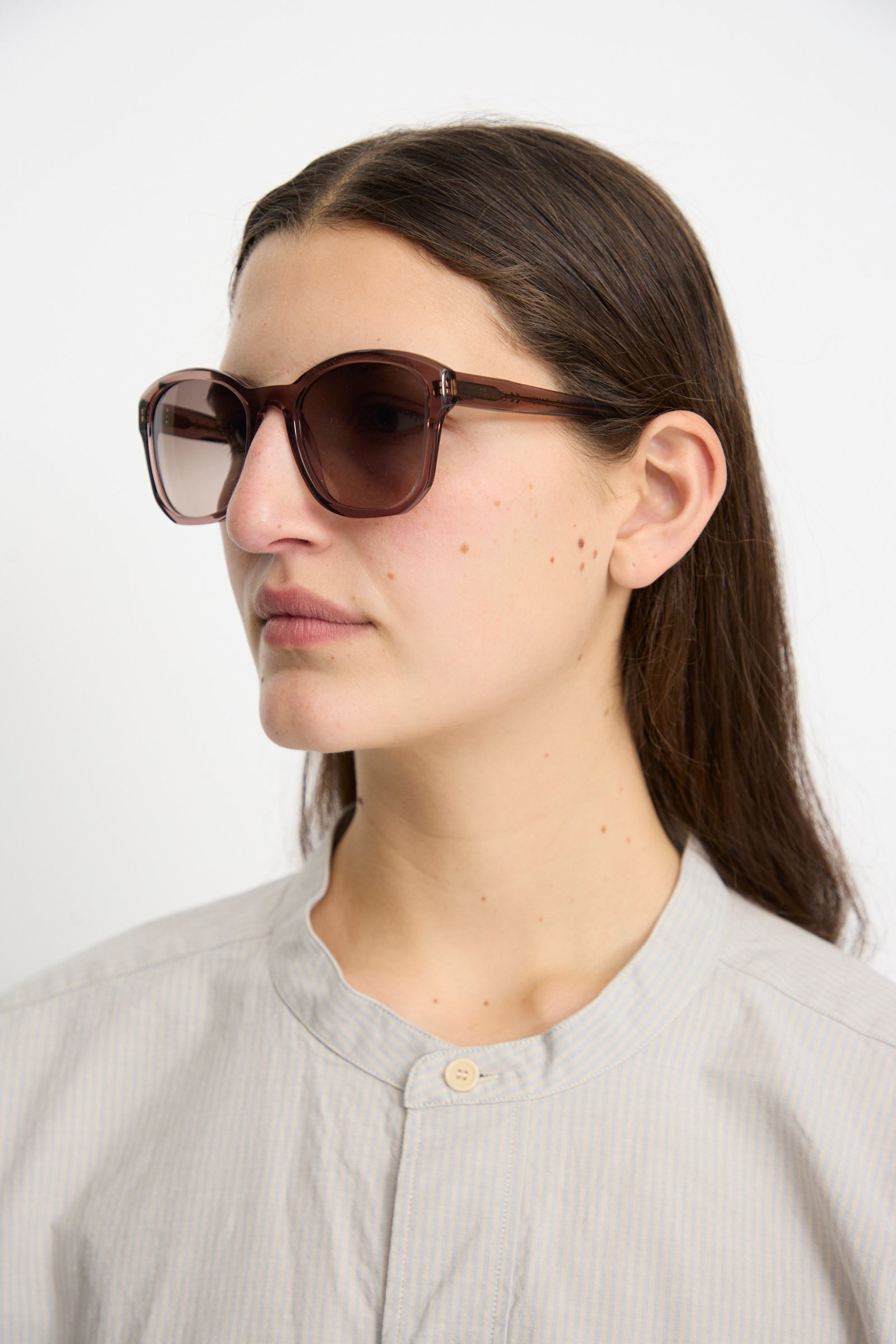 A person with long brown hair wears BQE Sunglasses in Bambi by Eva Masaki, featuring Zeiss lenses and a light gray buttoned shirt, glancing to the side against a plain background.