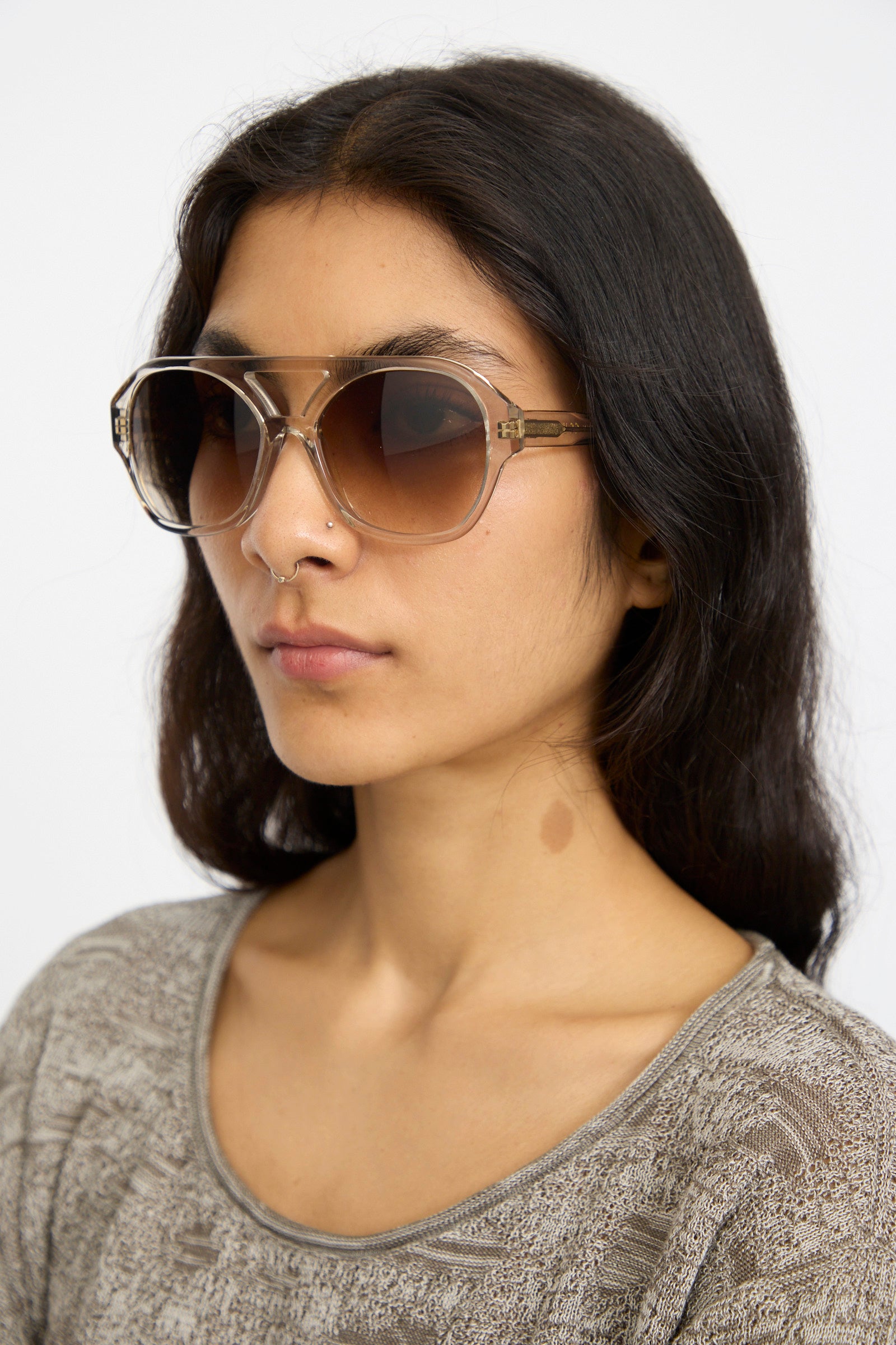 A woman with long dark hair wears Eva Masaki Chiyo Aviator Sunglasses in Hinoki and a patterned gray top, facing slightly to the side against a plain background.