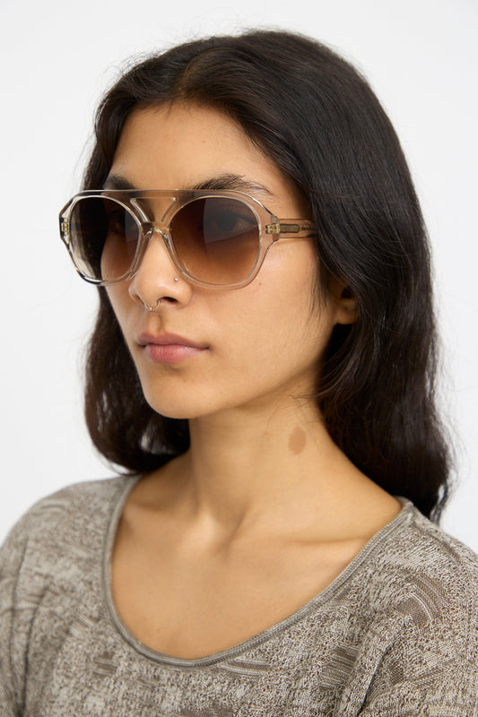 A woman with long dark hair wears Eva Masaki Chiyo Aviator Sunglasses in Hinoki and a patterned gray top, facing slightly to the side against a plain background.
