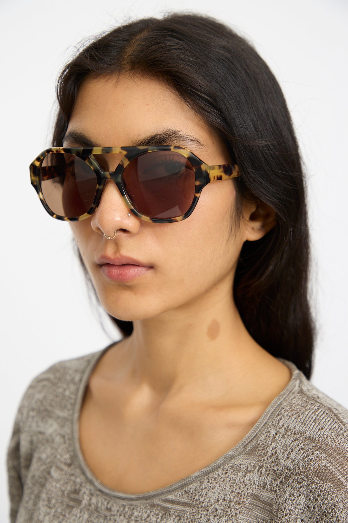 A person wearing Eva Masaki Chiyo Aviator Sunglasses in Tokyo and a textured light brown top, with long dark hair and a small nose ring, stands against a plain background.